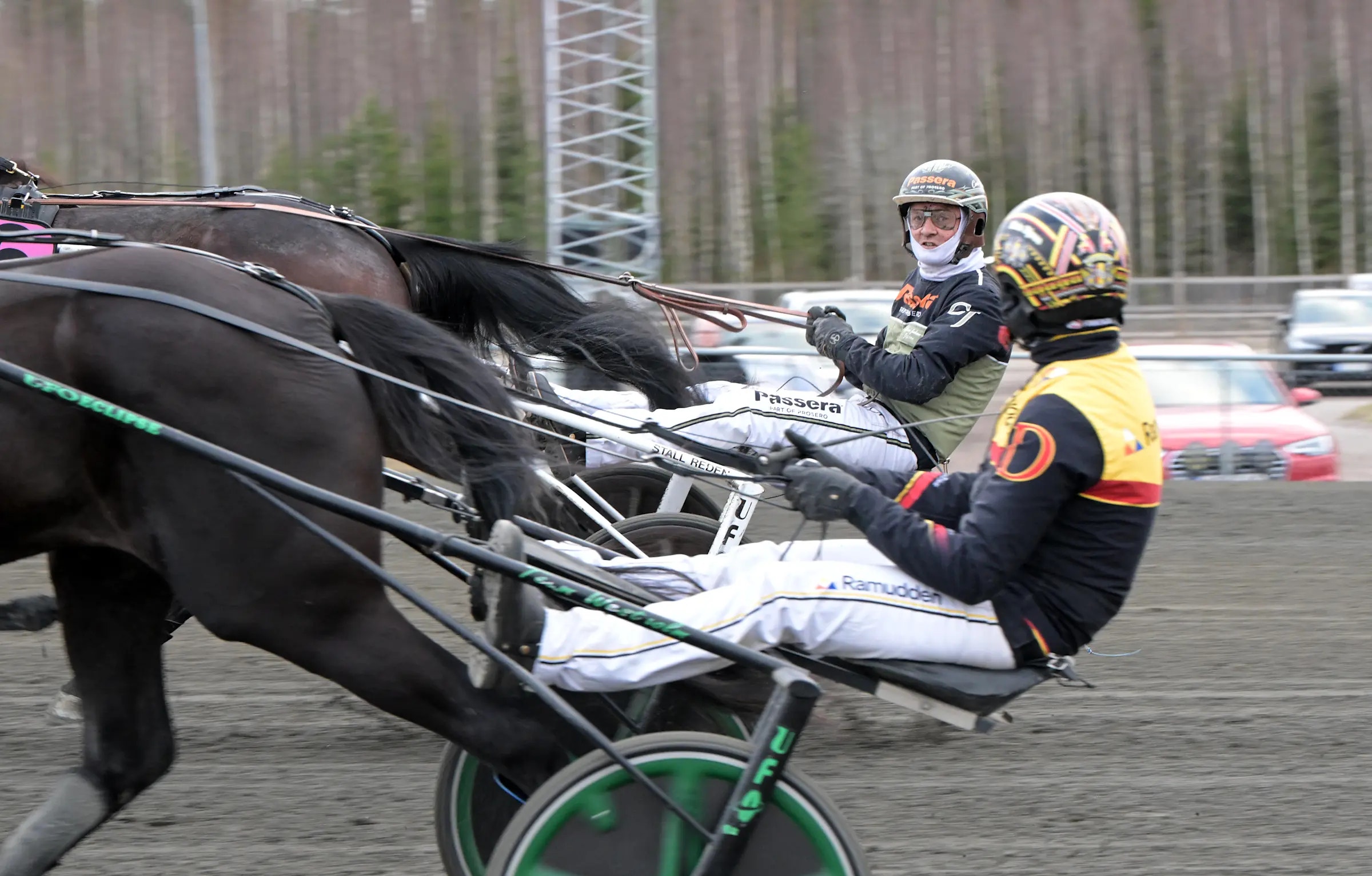 Två kuskar i travkusk-vagn blir dragna av en svart häst under ett travlopp.