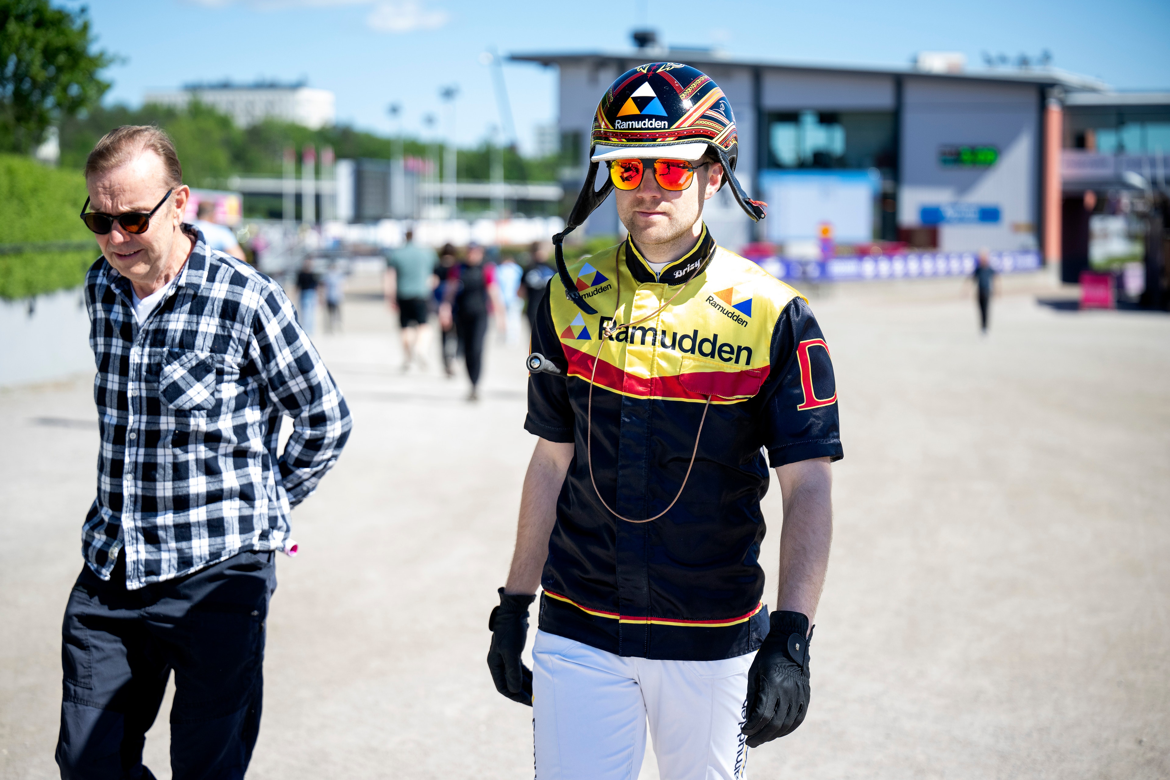 En cyklist i gul och svart tävlingsdräkt med cykelhjälm och solglasögon går tillsammans med en man i rutig skjorta på en eventplats.
