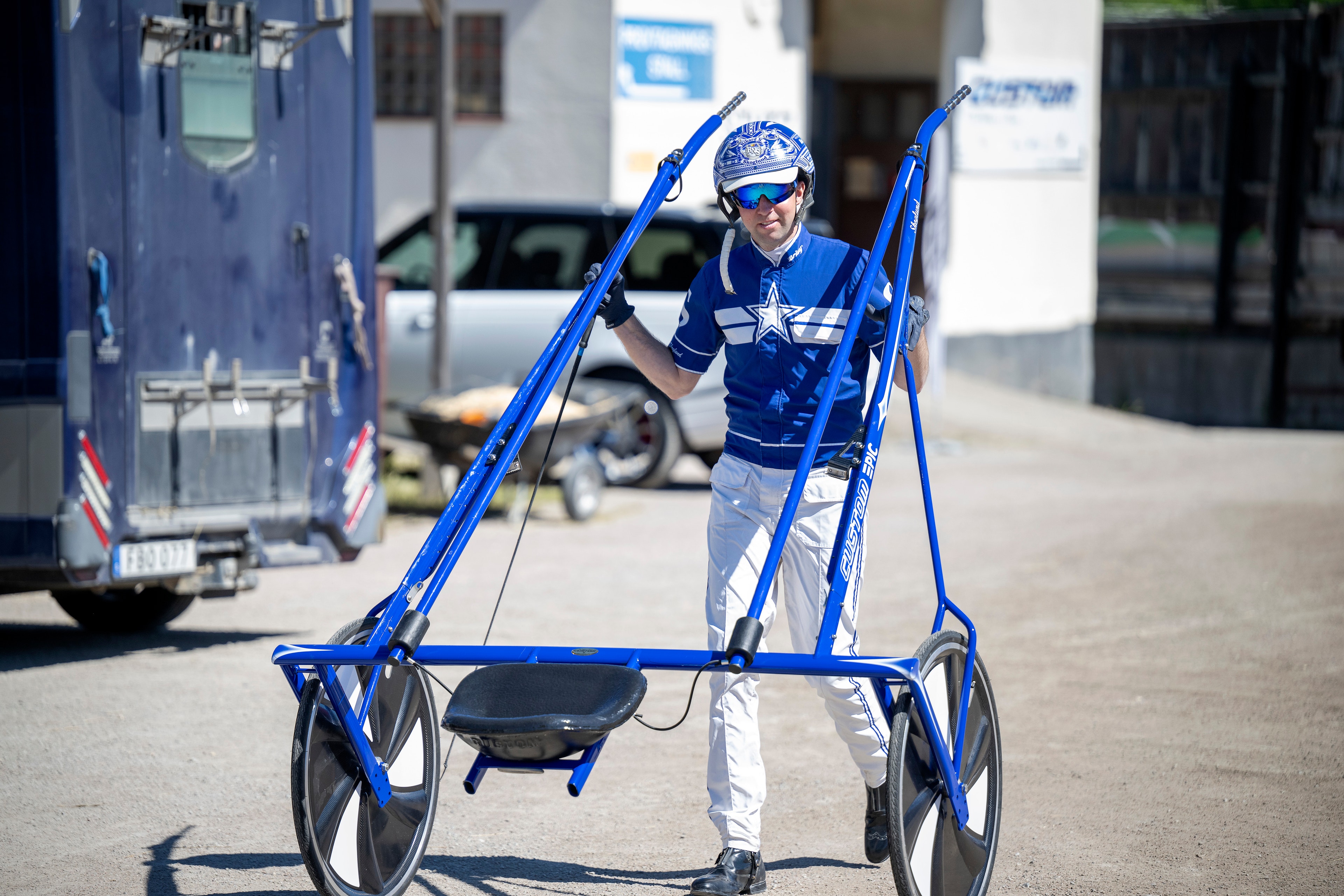 Person i blå skidkläder och hjälm håller upp ett par blå skidor ovanför ett blått skidåkningsfordon på en parkeringsplats.
