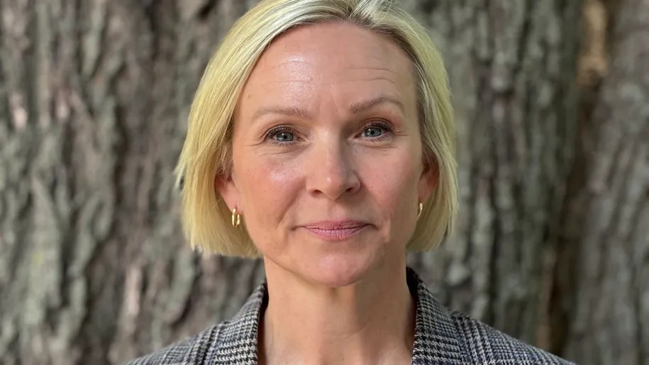 Porträtt av en kvinna med blont kort hår som ler mot kameran. Hon bär en grå kavaj och står framför en trädbark.