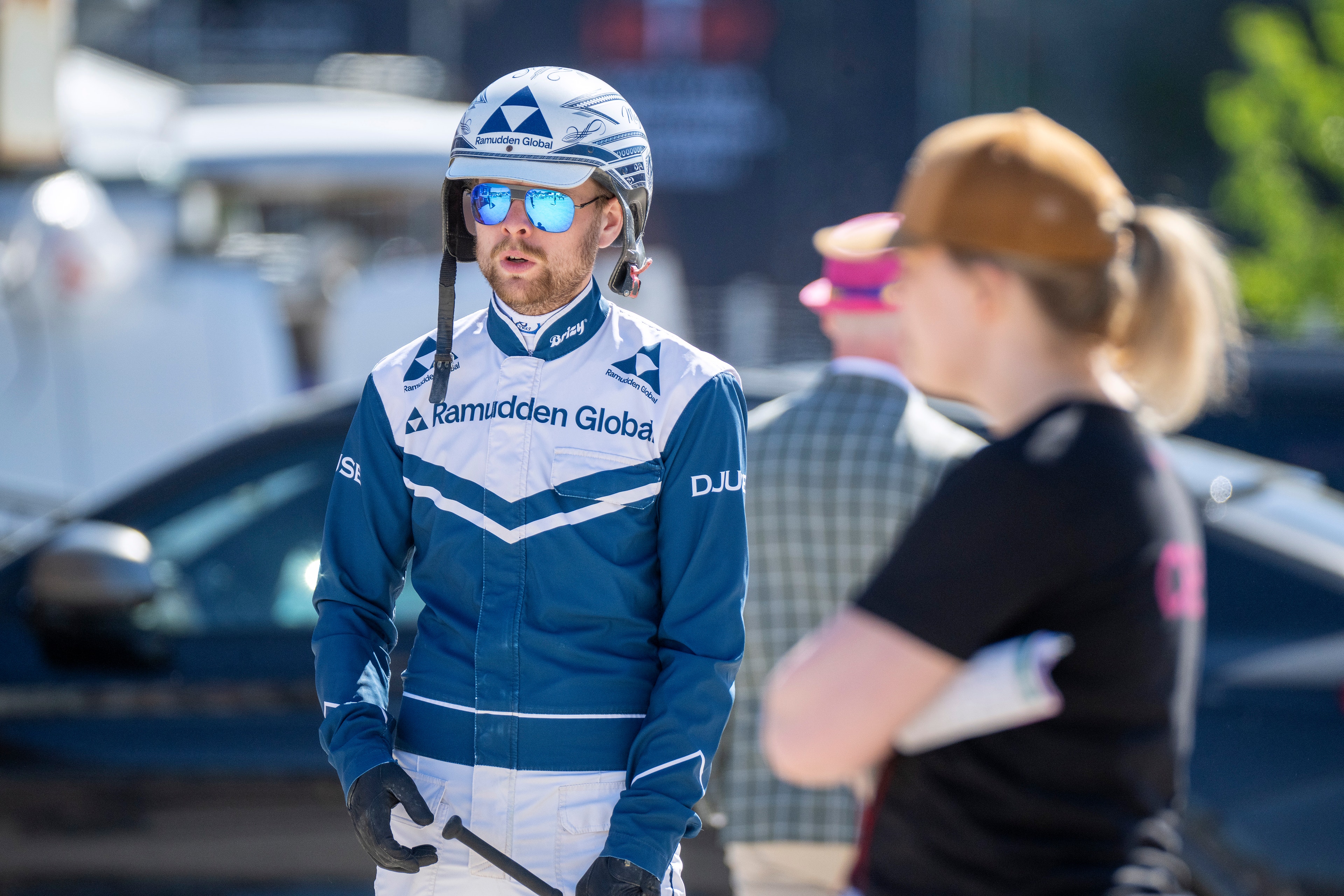 Cyklist i blå och vit tävlingsdräkt med hjälm och solglasögon står på en väg omgiven av andra personer.