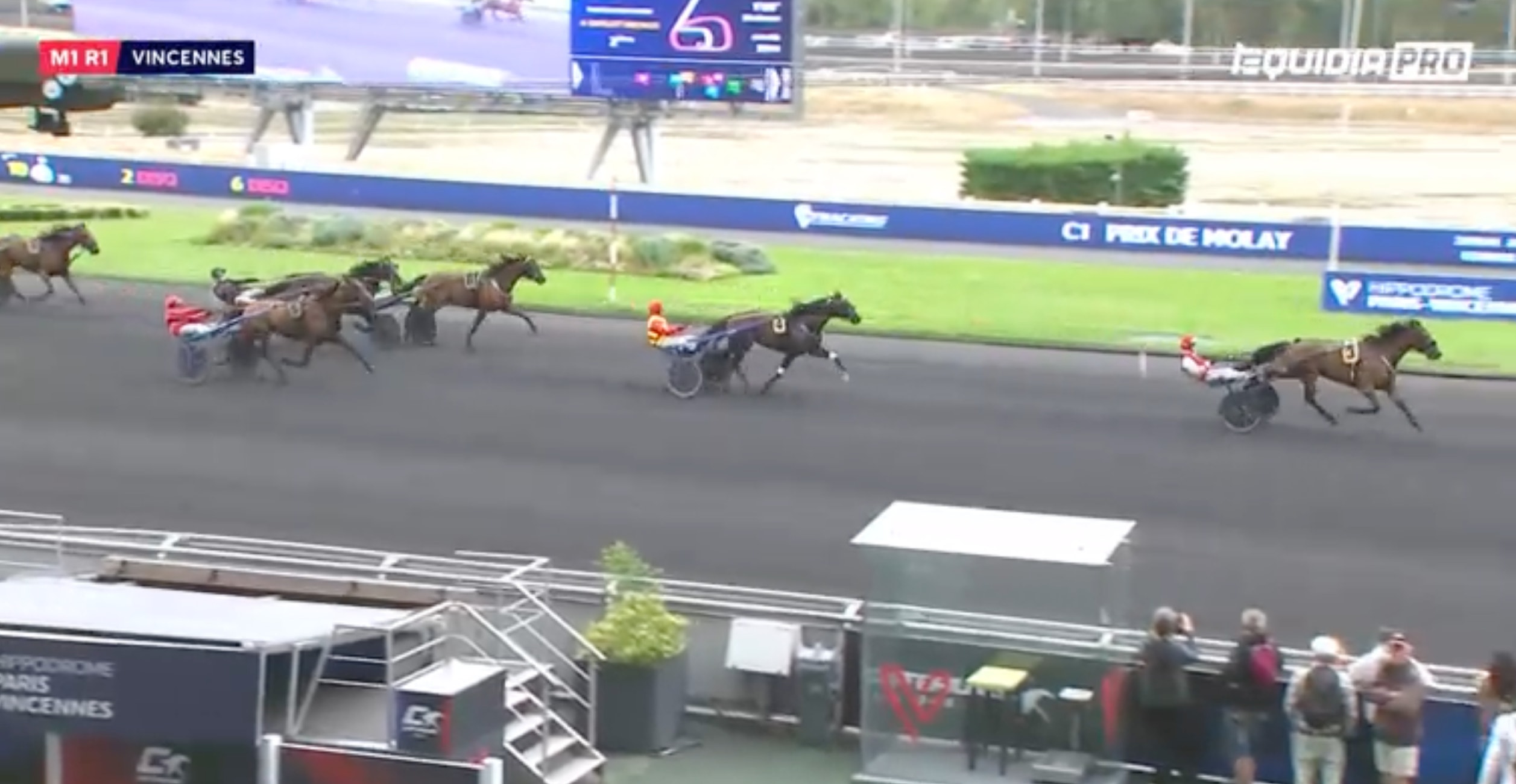 Hästkapplöpning på Vincennes med flera hästar och kuskar i sulky som tävlar på banan. Publiken sitter längs sidan och tittar på loppet.