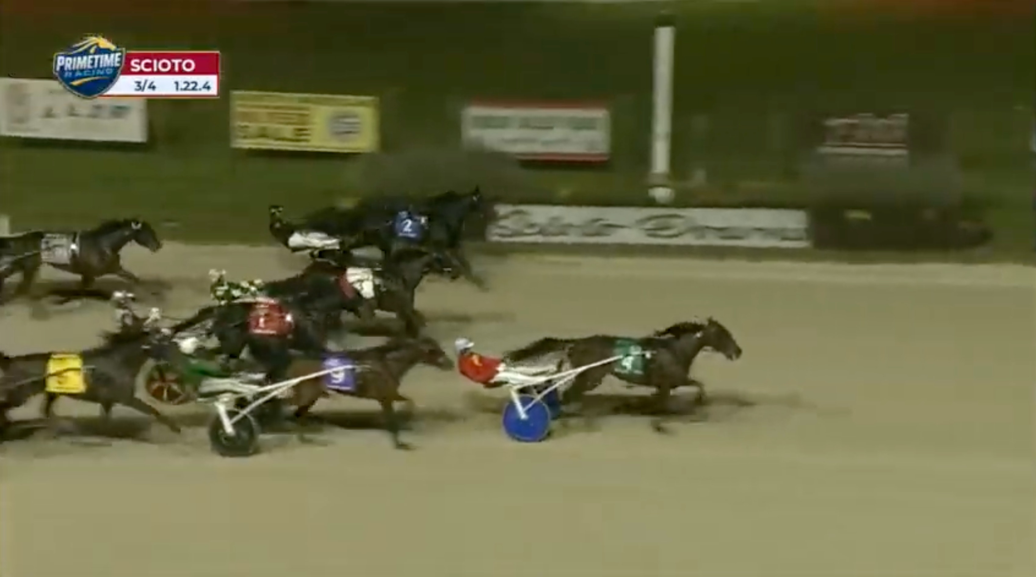Skärmbild från en travtävling på Scioto Downs. Flera hästar med förare i sulkys travar på en grusbana, och tävlingens tider visas överst i bilden.