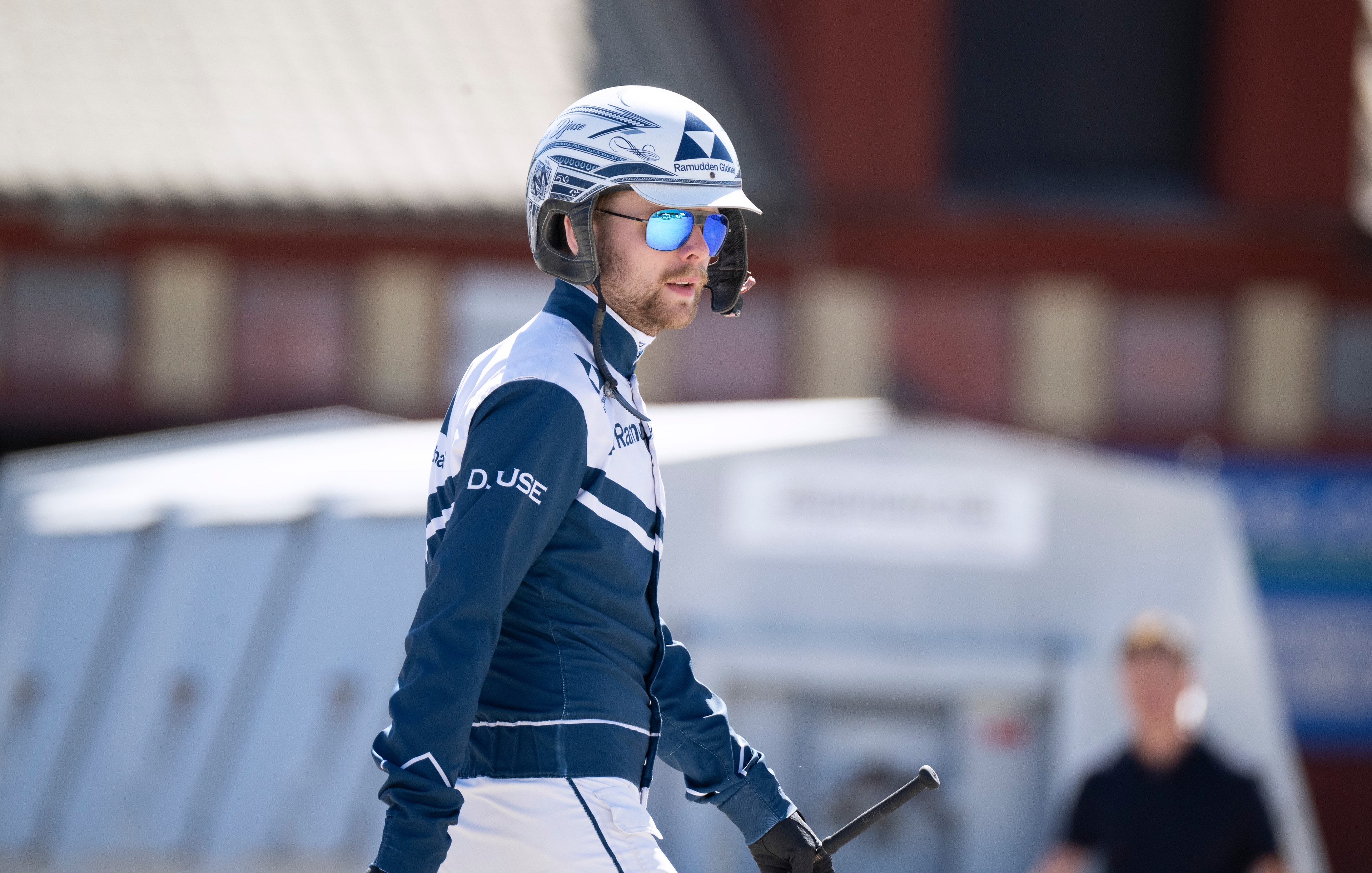 En cyklist i mörkblå och vit sportdräkt med hjälm och solglasögon håller sin cykel. I bakgrunden syns en vit tältstruktur och en rödbrun byggnad.