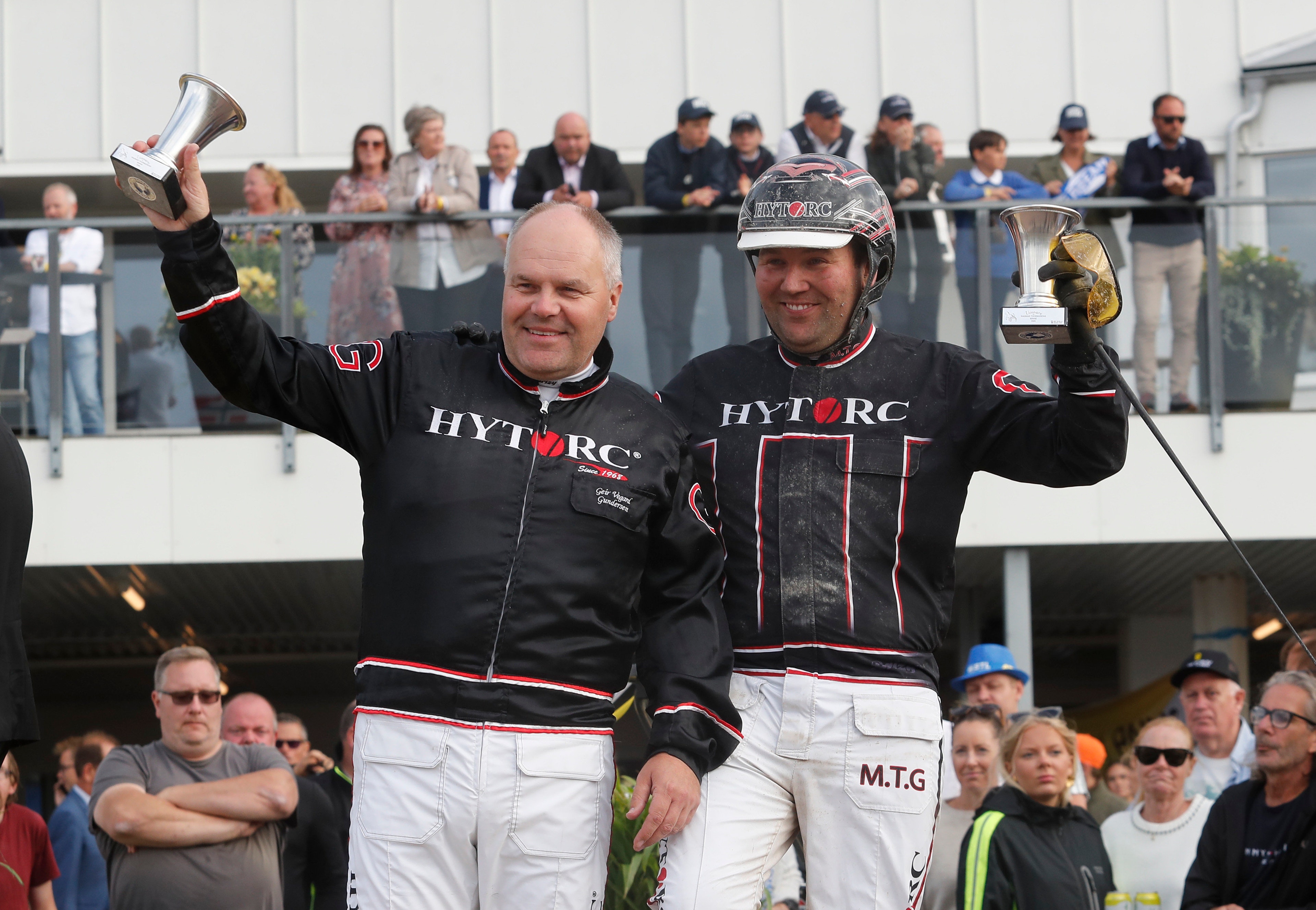Två racerförare på en vinnarpodium som höjer pokaler och trofeer i luften för att fira sin seger. De är klädda i svarta och vita HYTRC racing-dräkter. Publik syns i bakgrunden.