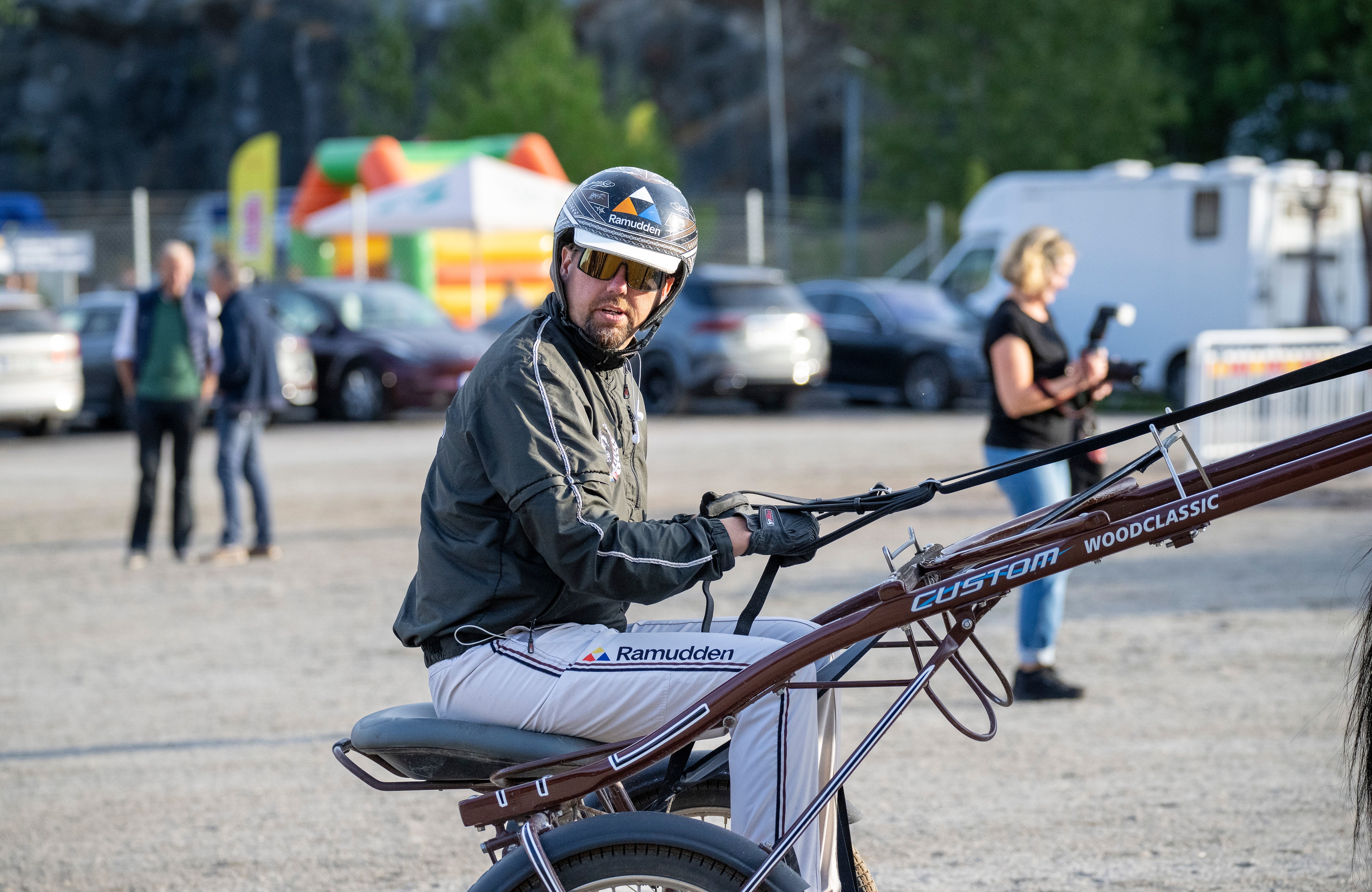 En man med cykelhjälm sitter på en cykel vid ett utomhusarrangemang. I bakgrunden syns parkeringsplats, andra människor och ett lekland.