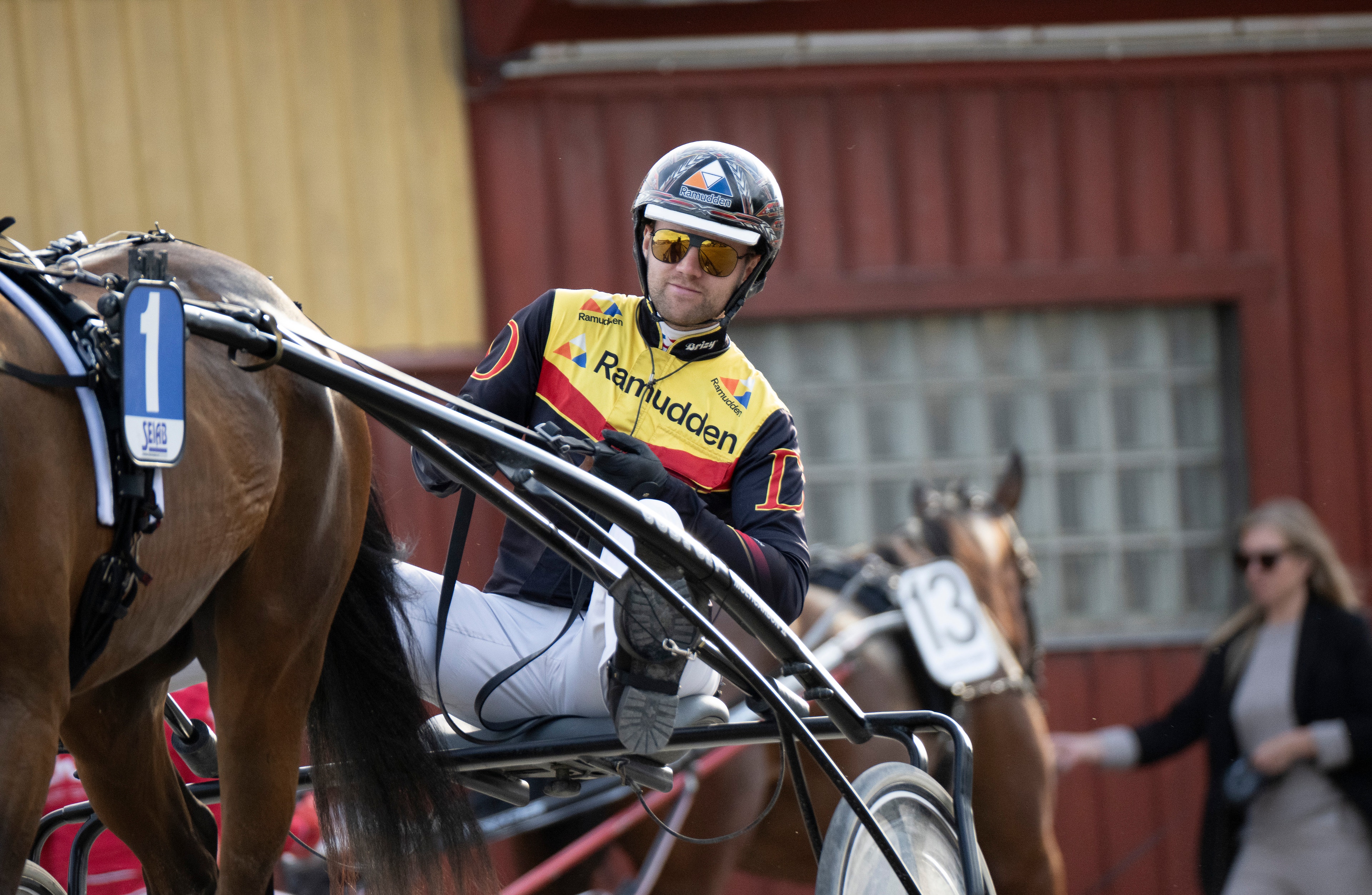 Kusken sitter i en sulky bakom en häst under ett travlopp. Kusken bär hjälm, solglasögon och en gul och röd tävlingsdräkt. Hästen är brunfärgad och är försedd med travutrustning. I bakgrunden syns en röd ladugård och några åskådare.