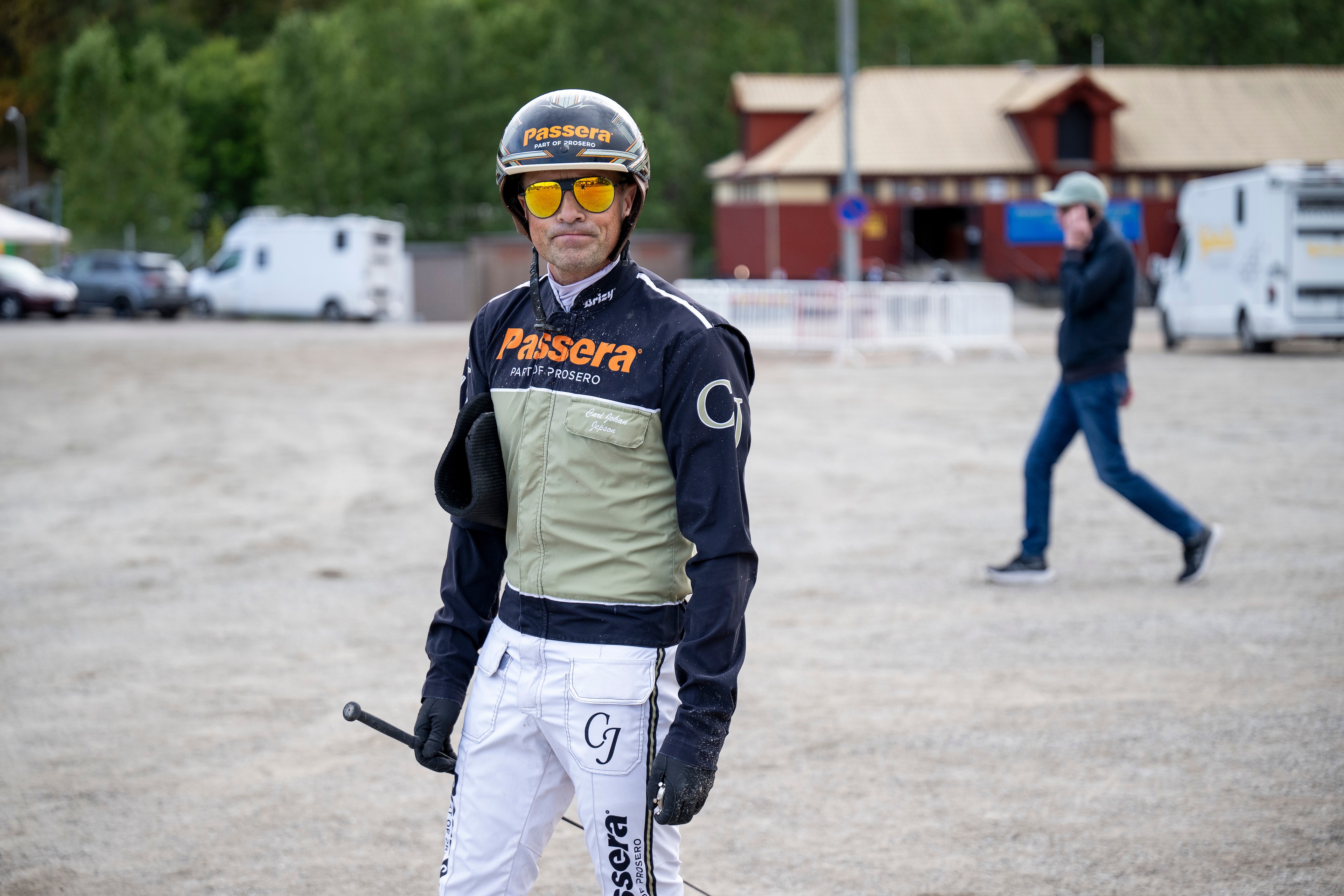 En ryttare i svart och beige ridkläder med gul hjälm och gula glasögon står på en ridarena med stall och parkeringsplats i bakgrunden.