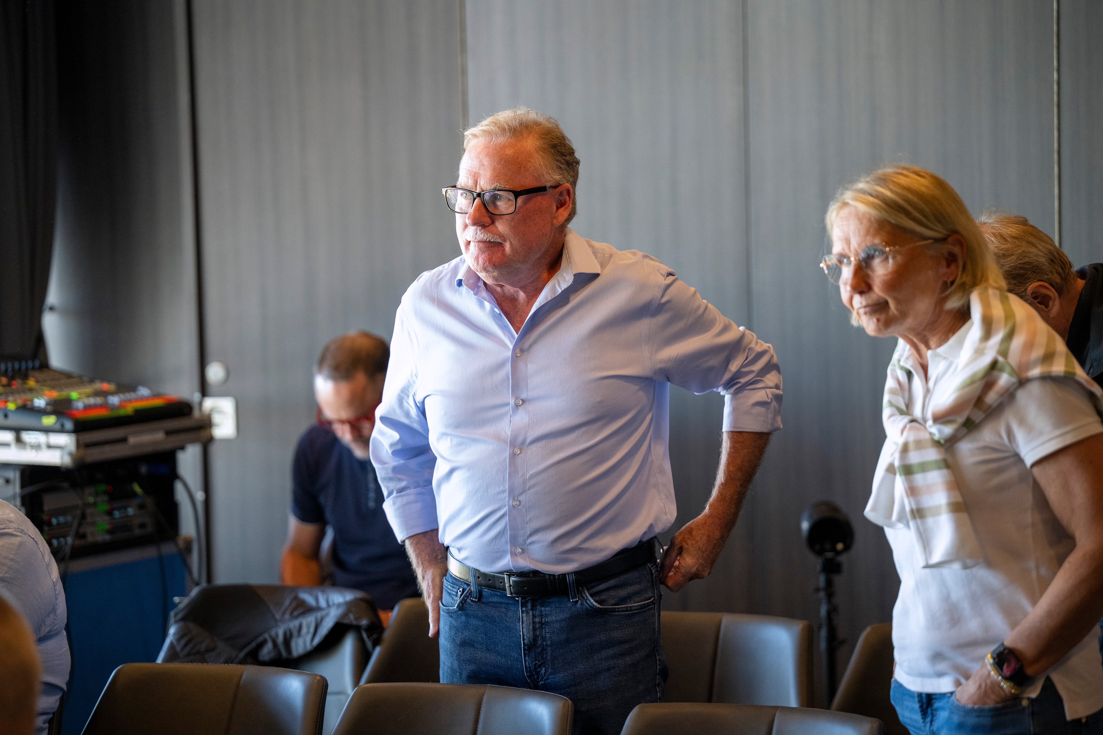 En man med glasögon i blå skjorta och en kvinna i vit skjorta står i ett konferensrum tillsammans med andra personer. De verkar vara på ett möte eller presentationstillfälle.