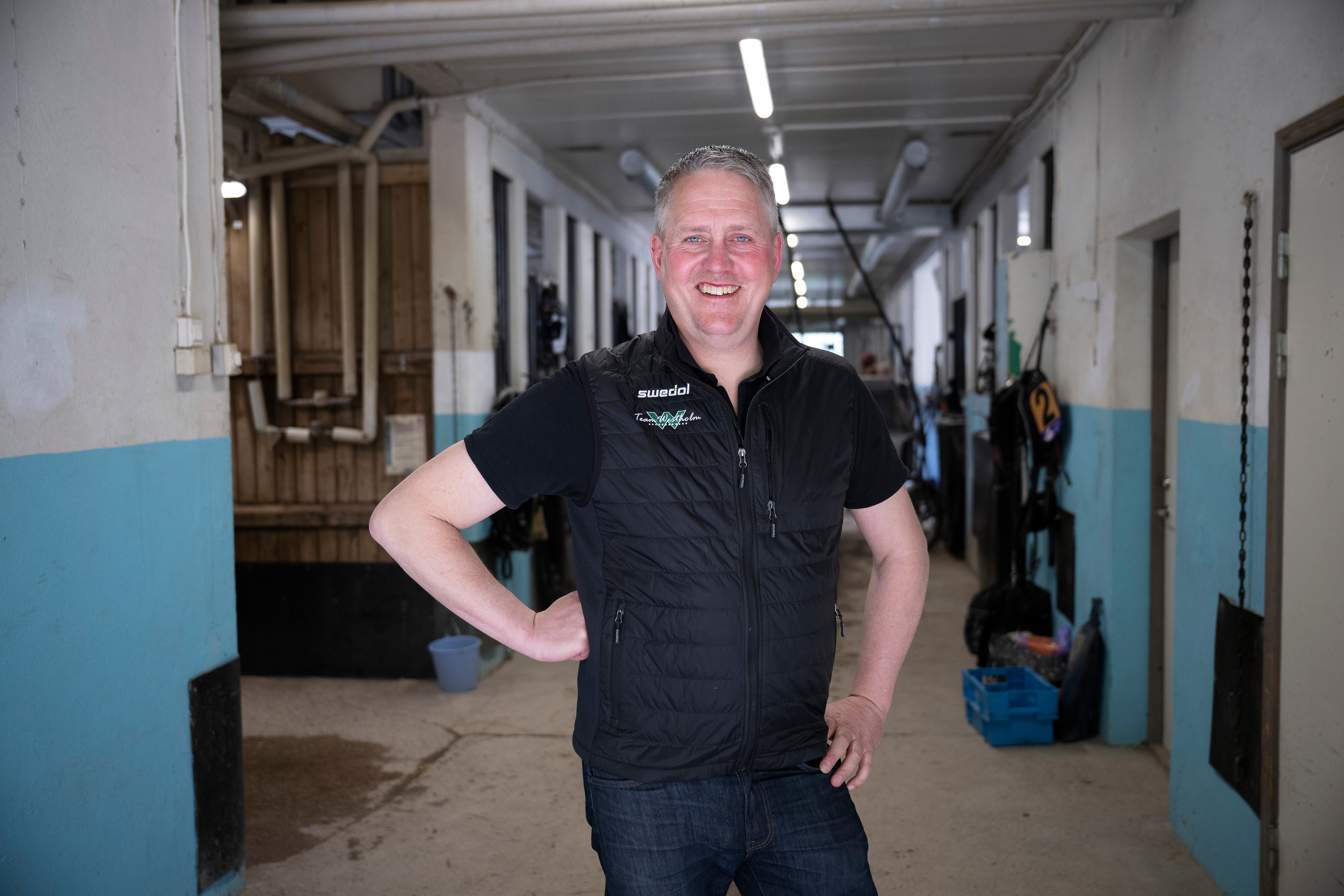 Porträttfoto av en man som står i en stall och ler mot kameran. Han är klädd i svart väst över svart t-shirt och jeans.