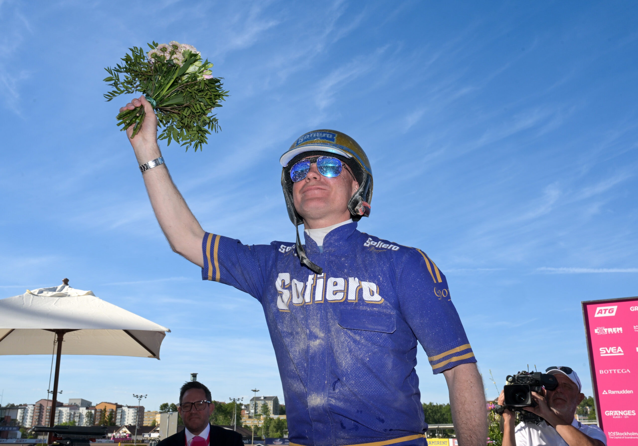 Cyklist i blå tröja håller upp en vinnarbukkett med gröna växter mot en blå himmel. Cyklisten bär solglasögon och en cykelhjälm.