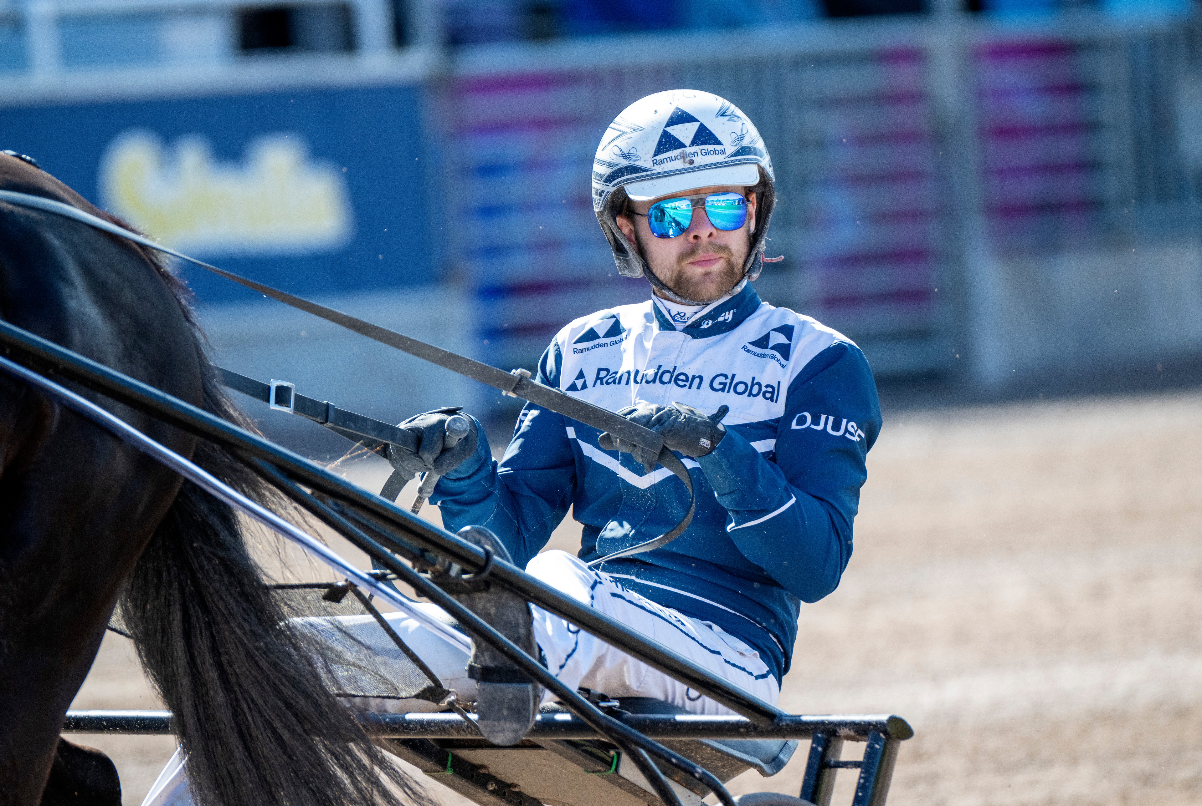 Kuskvagnskörare i blå och vit tävlingsdräkt sitter i en vagn bakom en häst vid Solvalla travbana.