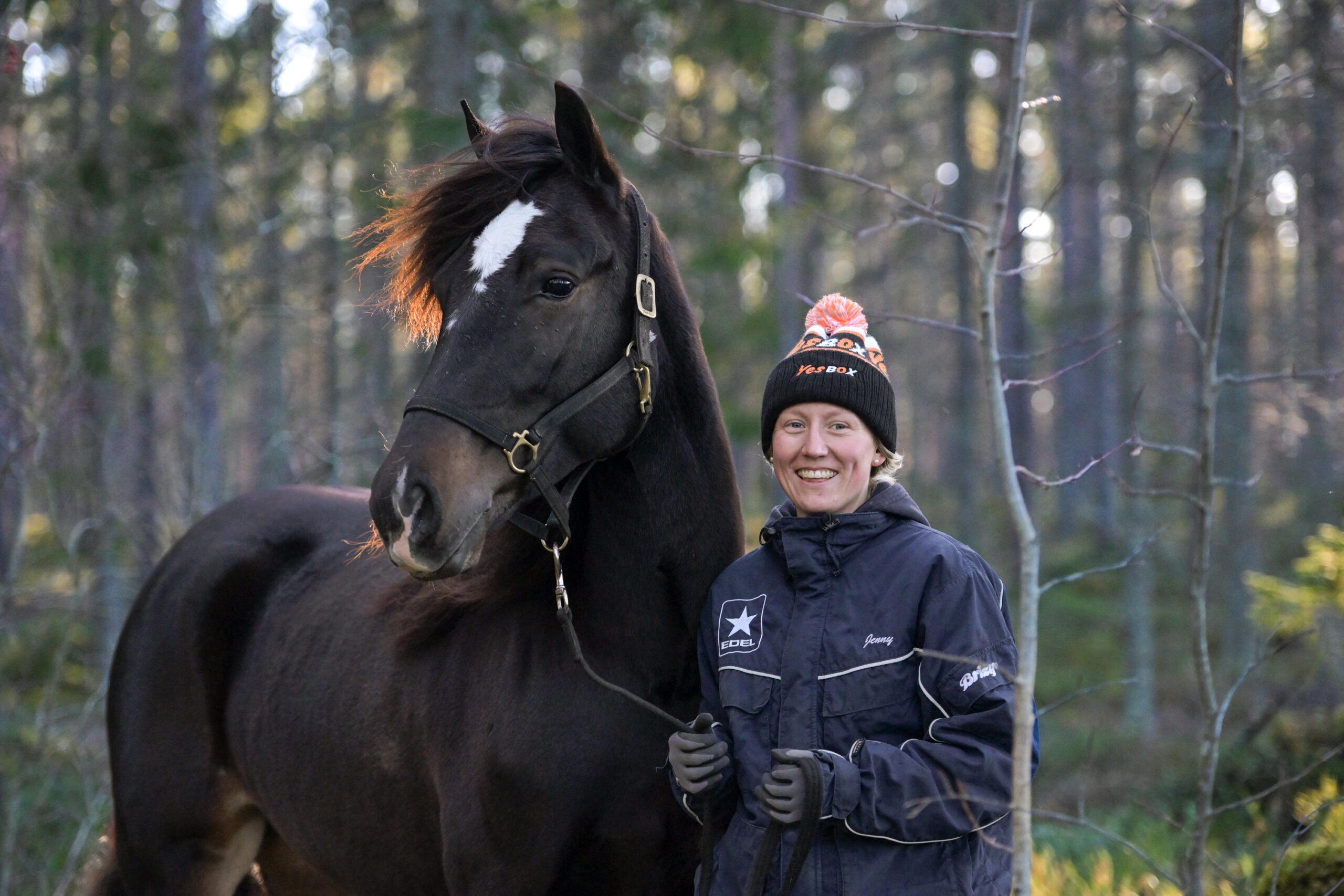 En person står tillsammans med en svart häst i skogen. Personen bär mössa och mörk jacka, och hästen har en vit stjärna på pannan.