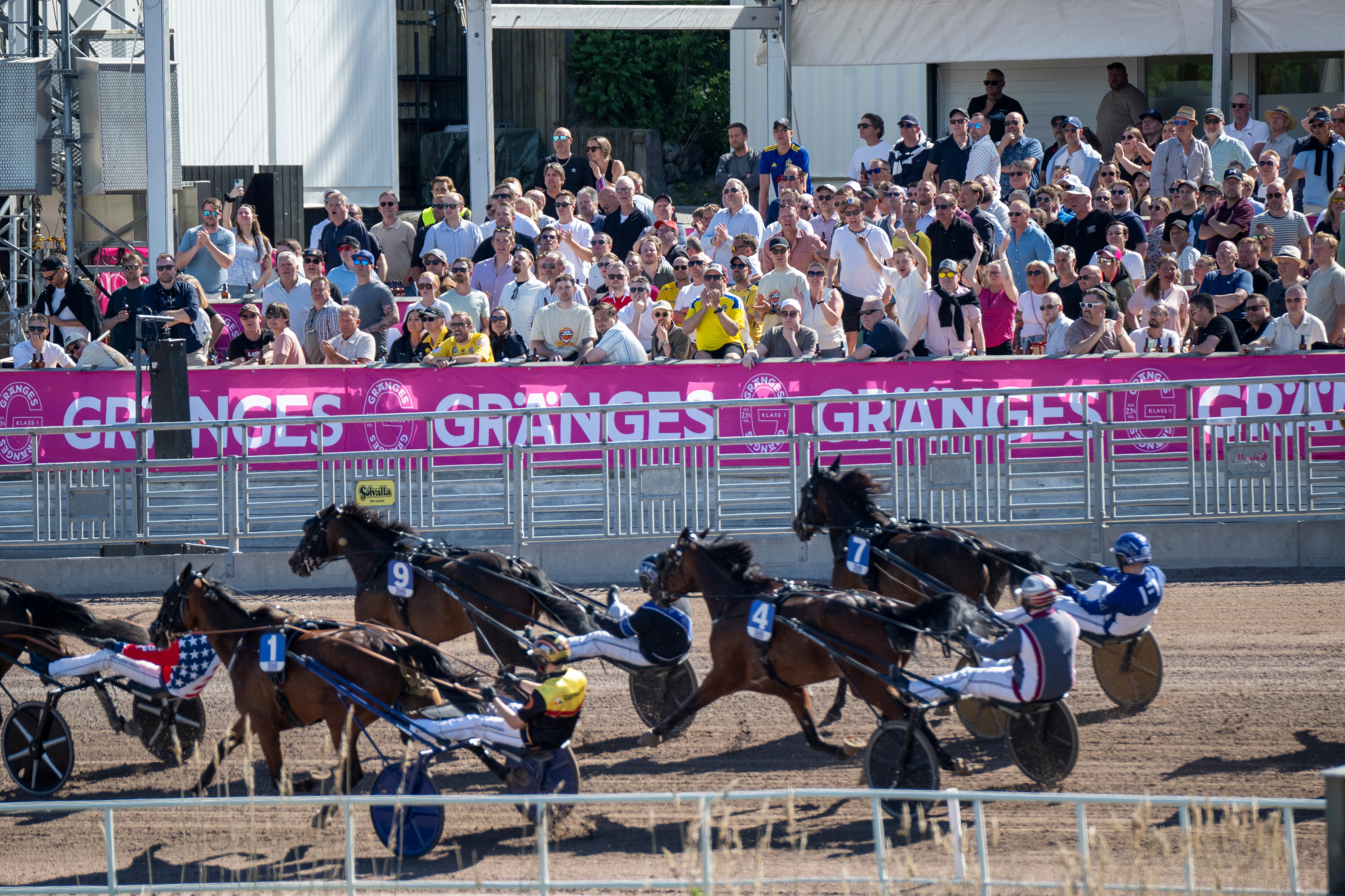 Travtävling på Solvalla med flera hästar och kuskar i tävlingsbanan. Publik sitter längs sidlinjen bakom rosa staket med Granges logotyp.