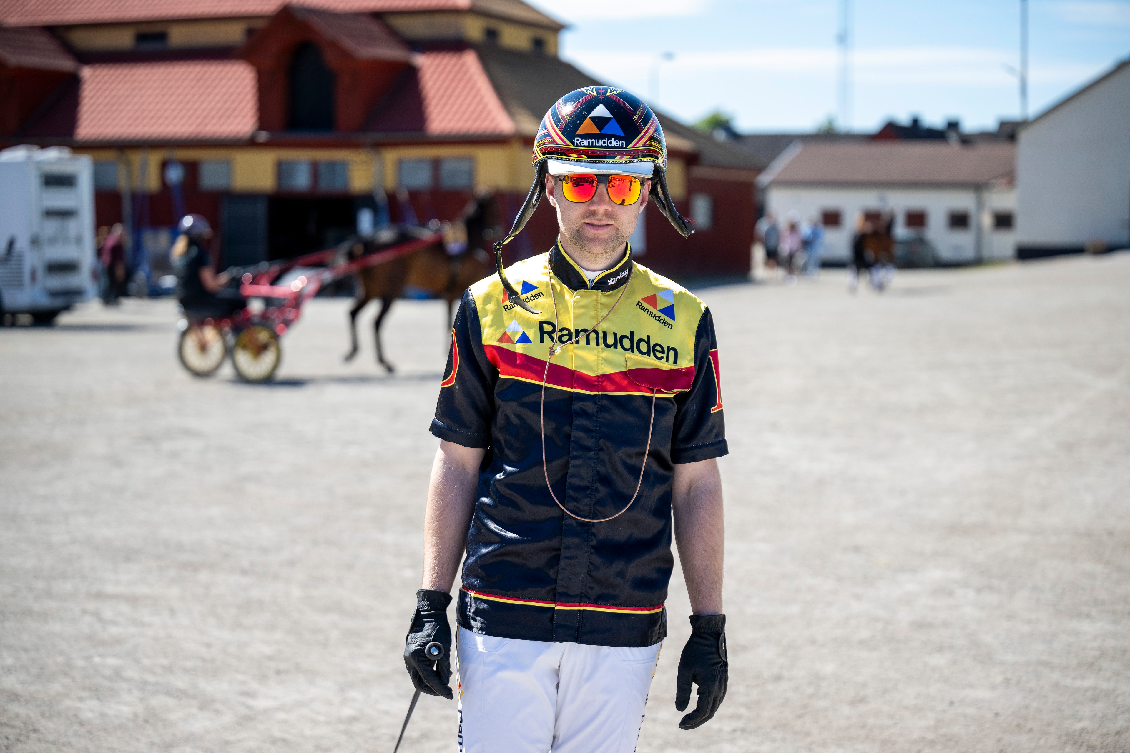 Porträttbild av en cyklist i gul och svart väst med texten Ramudden, vita cykelbyxor och cykelhjälm. I bakgrunden syns travhästar och byggnader på Solvalla travbana.