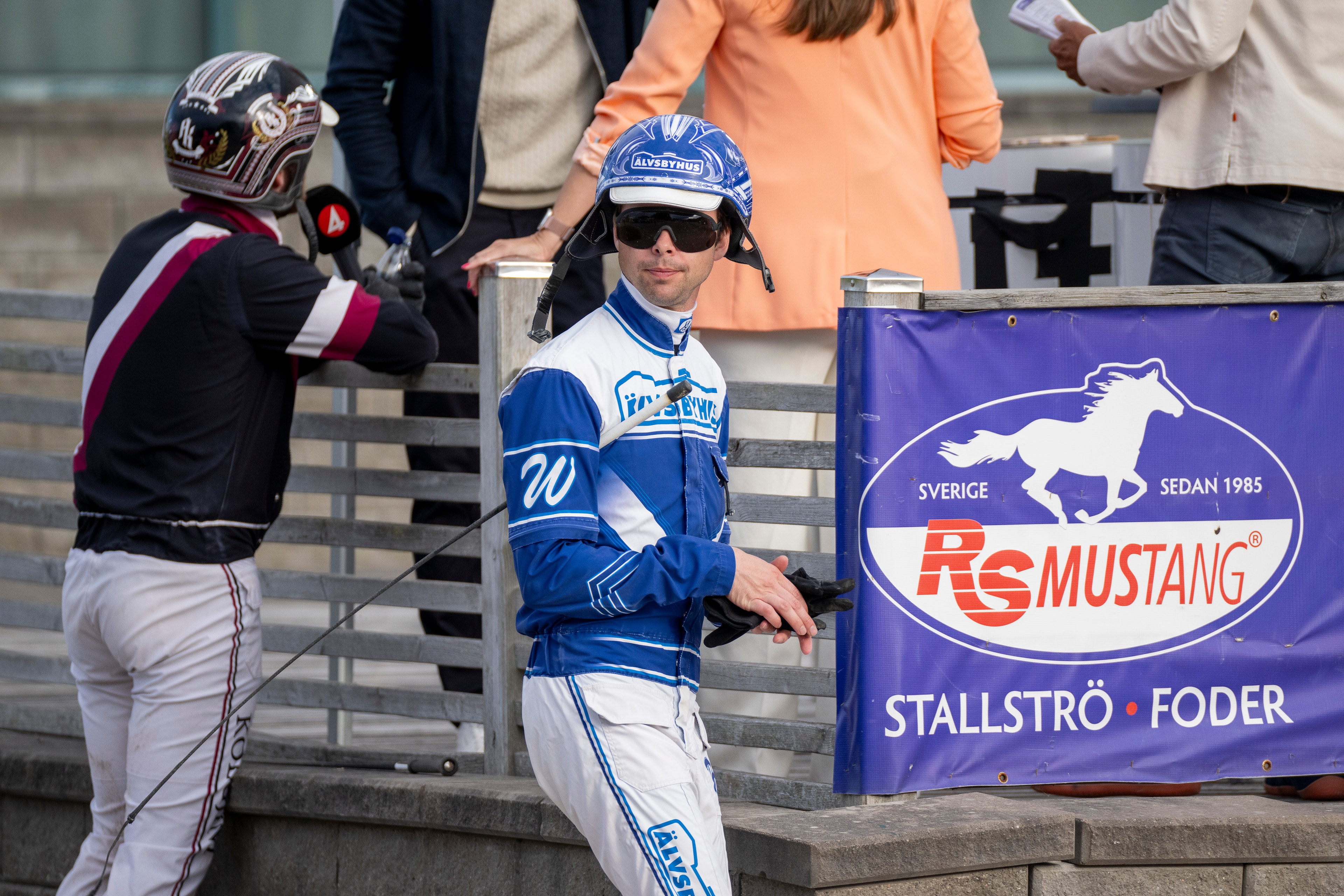 Motorcykelförare i blå racingkläder och hjälm står vid en racingbana tillsammans med andra personer. En bannerbräda med RS Mustang-logon och texten 'Sverige sedan 1985 Stallströfoder' syns i bakgrunden.
