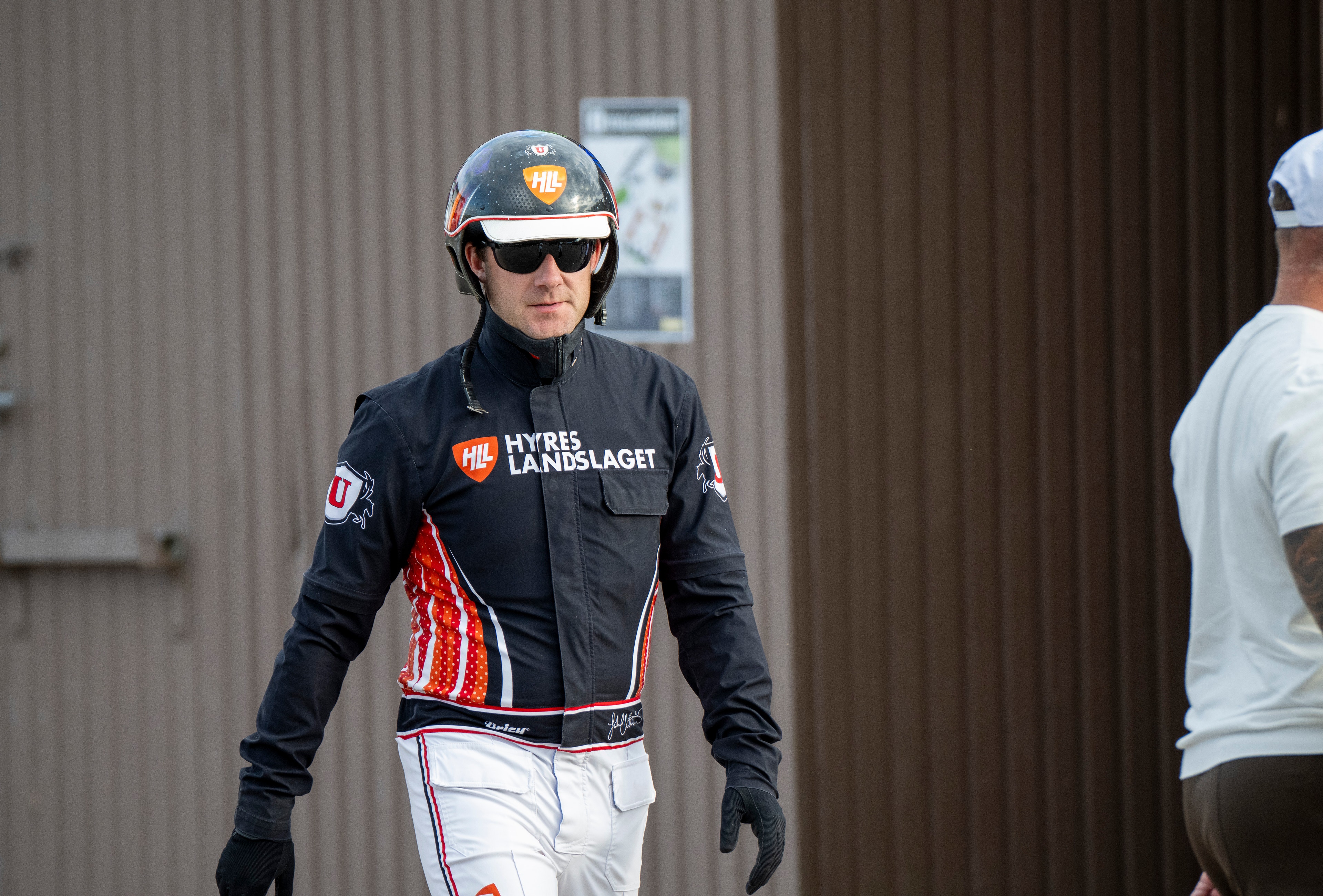 En person i svart racingdräkt med hjälm och solglasögon går framför en korrugerad metallvägg.