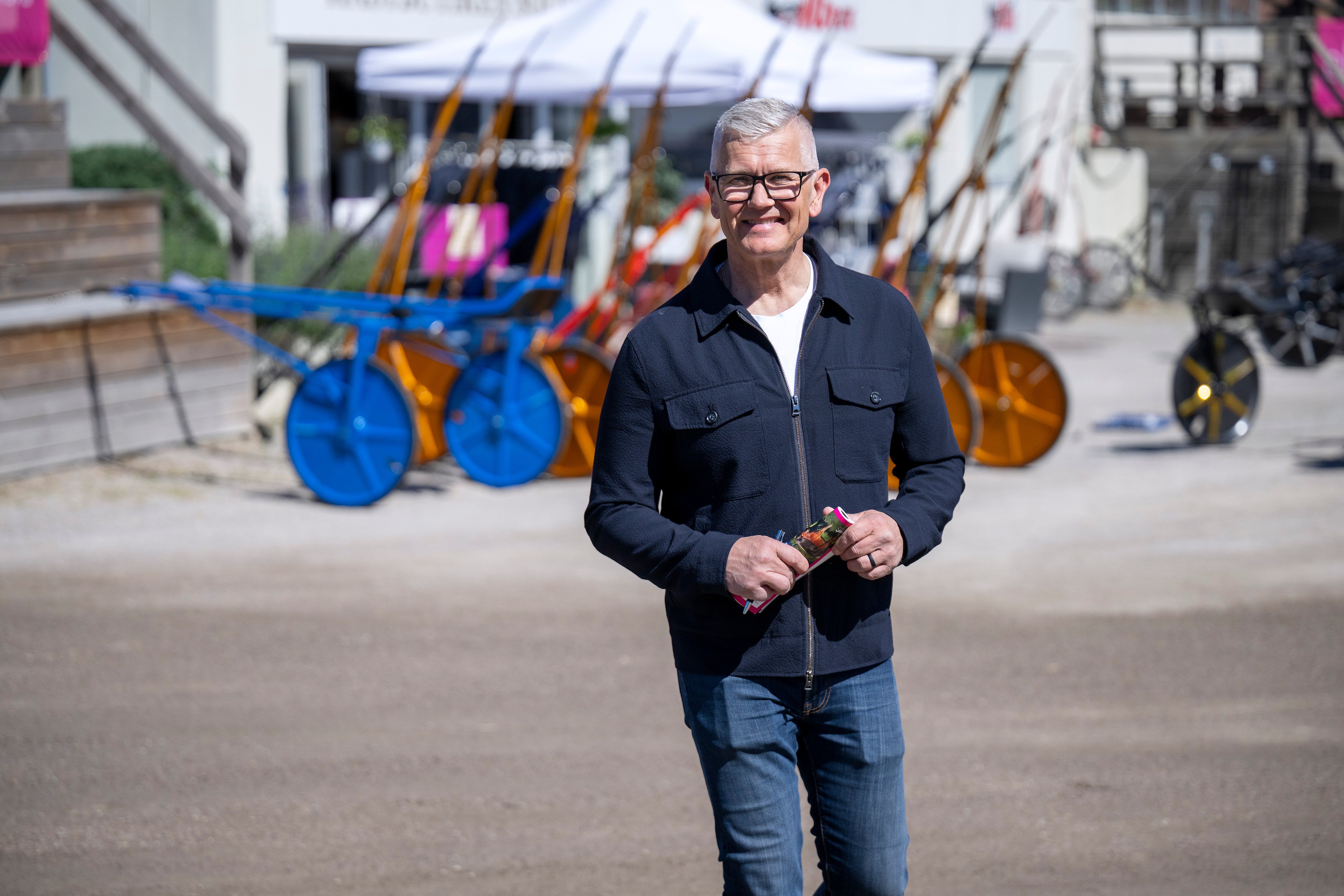 Porträtt av en äldre man med glasögon som står utomhus på en gård. Han bär en mörk skjorta och jeans. I bakgrunden syns färgglad utrustning och maskiner.