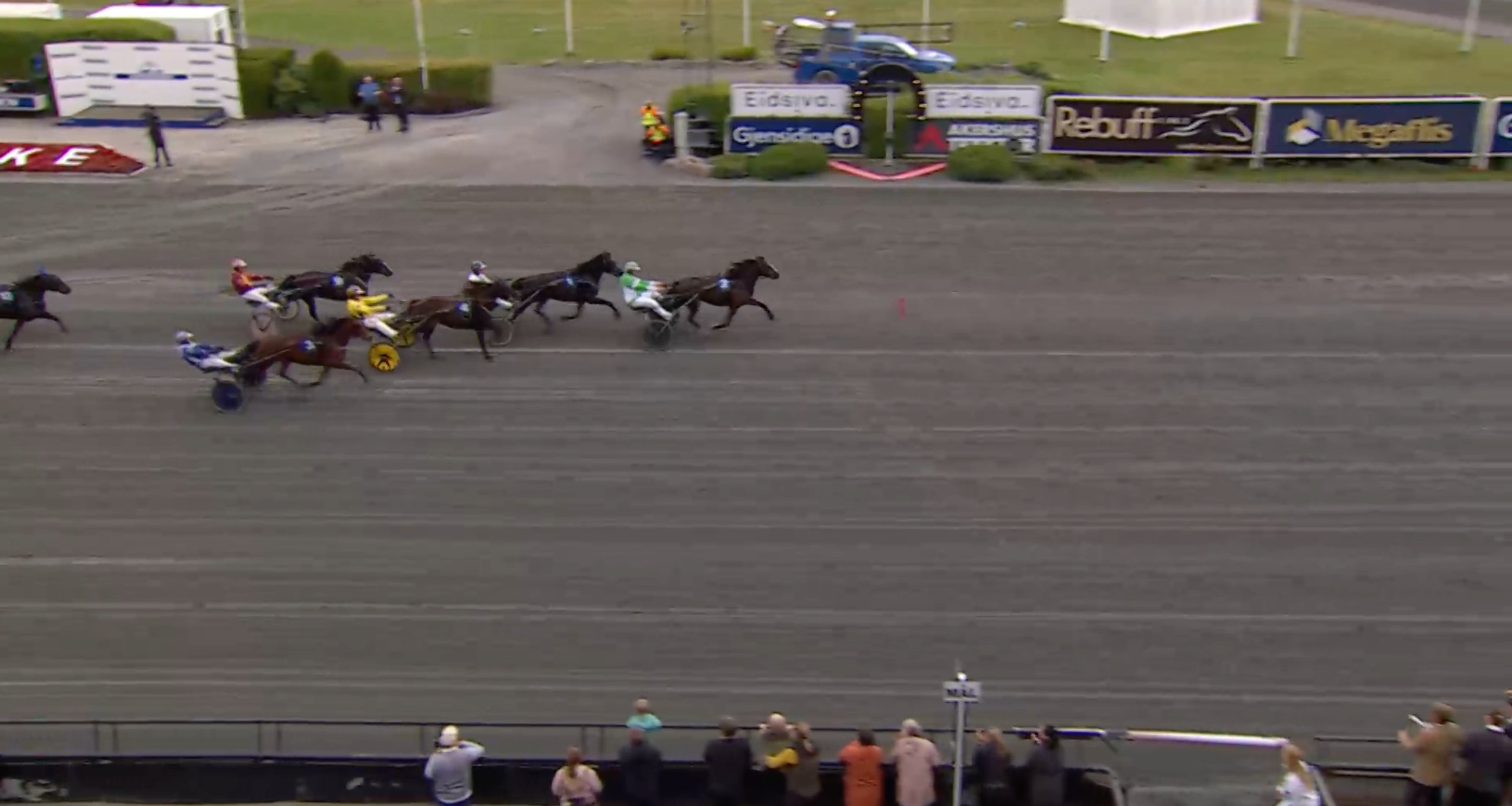 Flera travhästar med sulky och kusk tävlar mot varandra på en travbana.