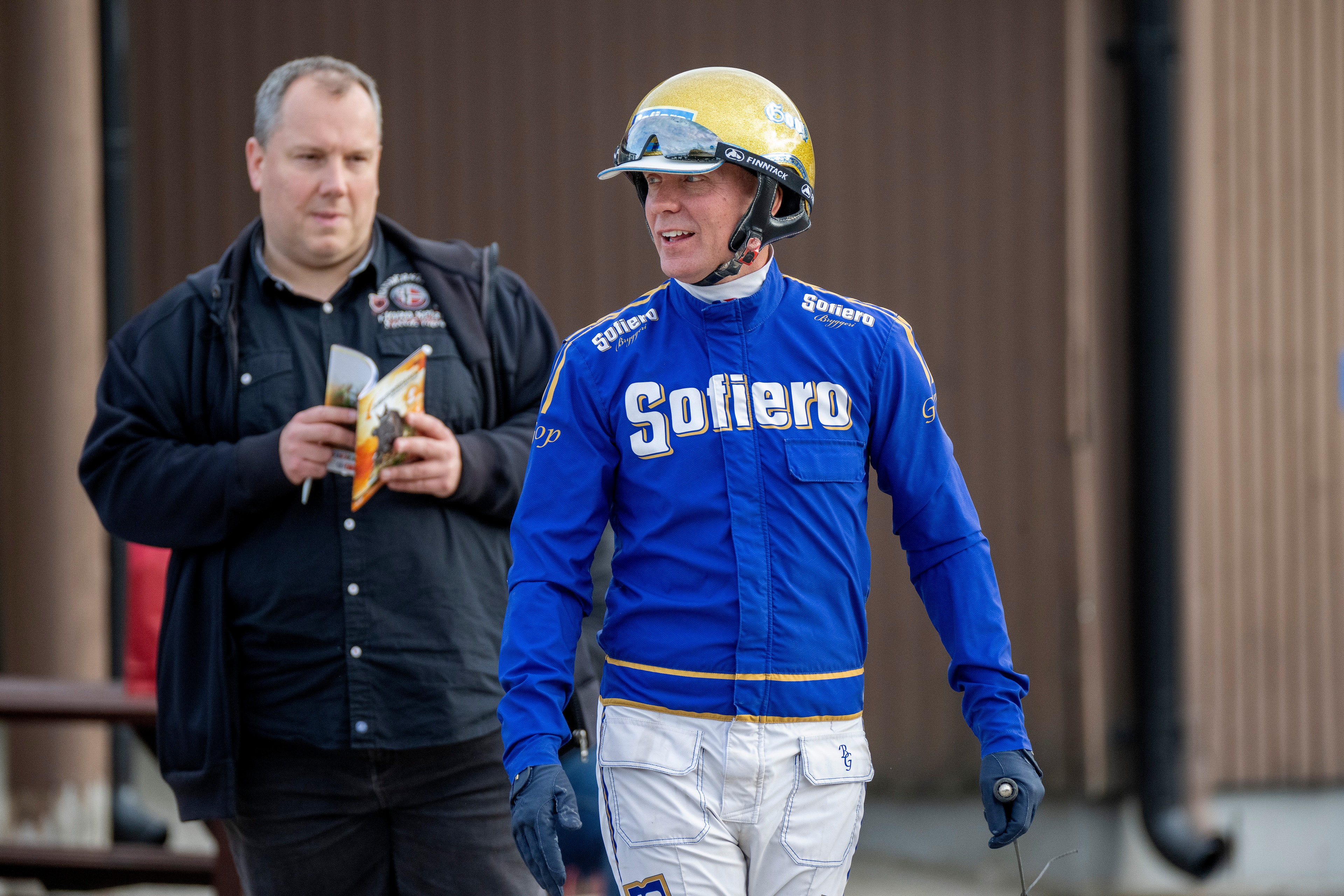 En racerförare i blå racingdräkt med gul hjälm tillsammans med en man i svart jacka som håller en banan. De står på en racerbana.
