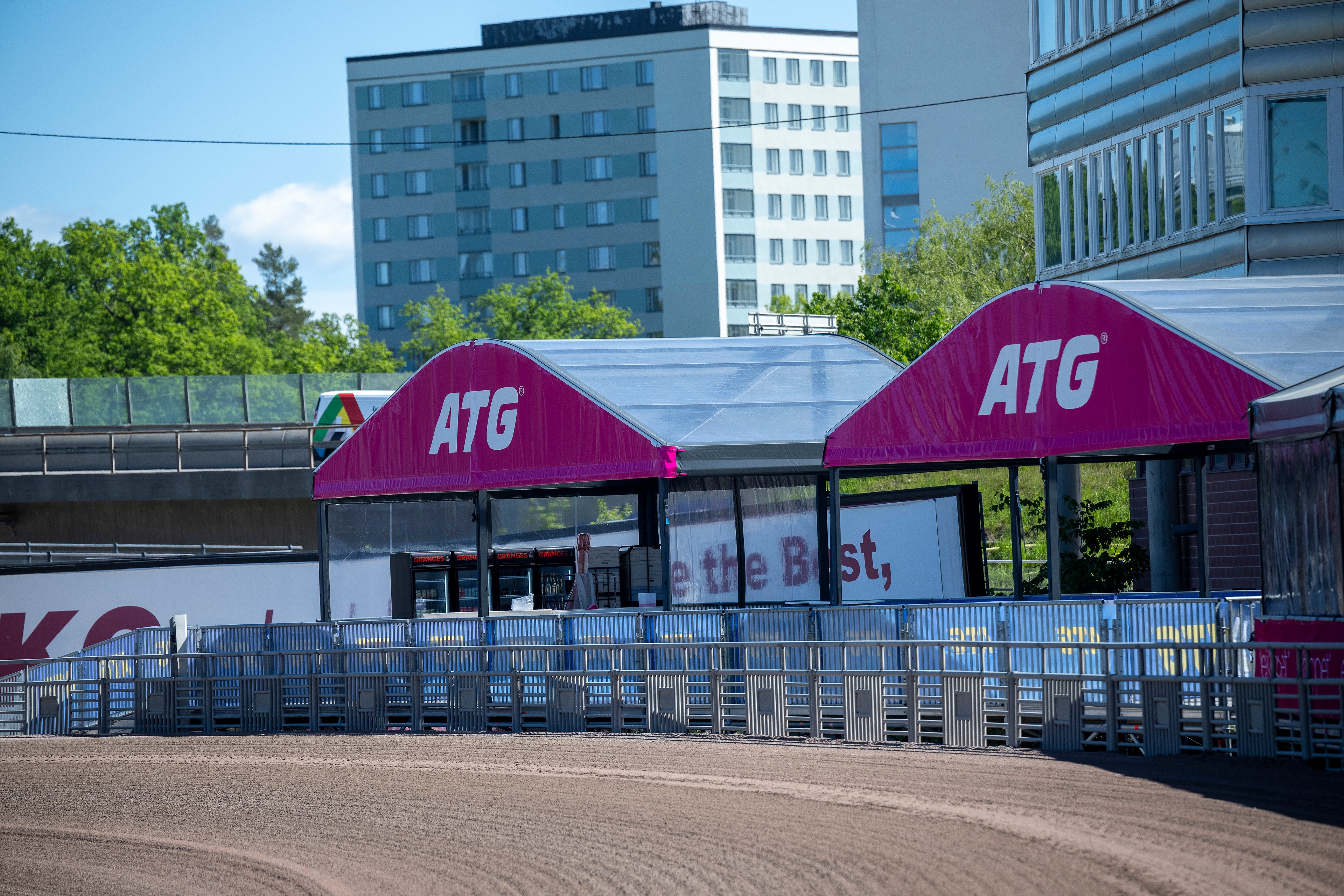 ATG-byggnad med magentafärgade tak vid en travbana. I förgrunden syns en sandbana och blå metallräcken. Moderna bostadshus syns i bakgrunden under en solig dag.