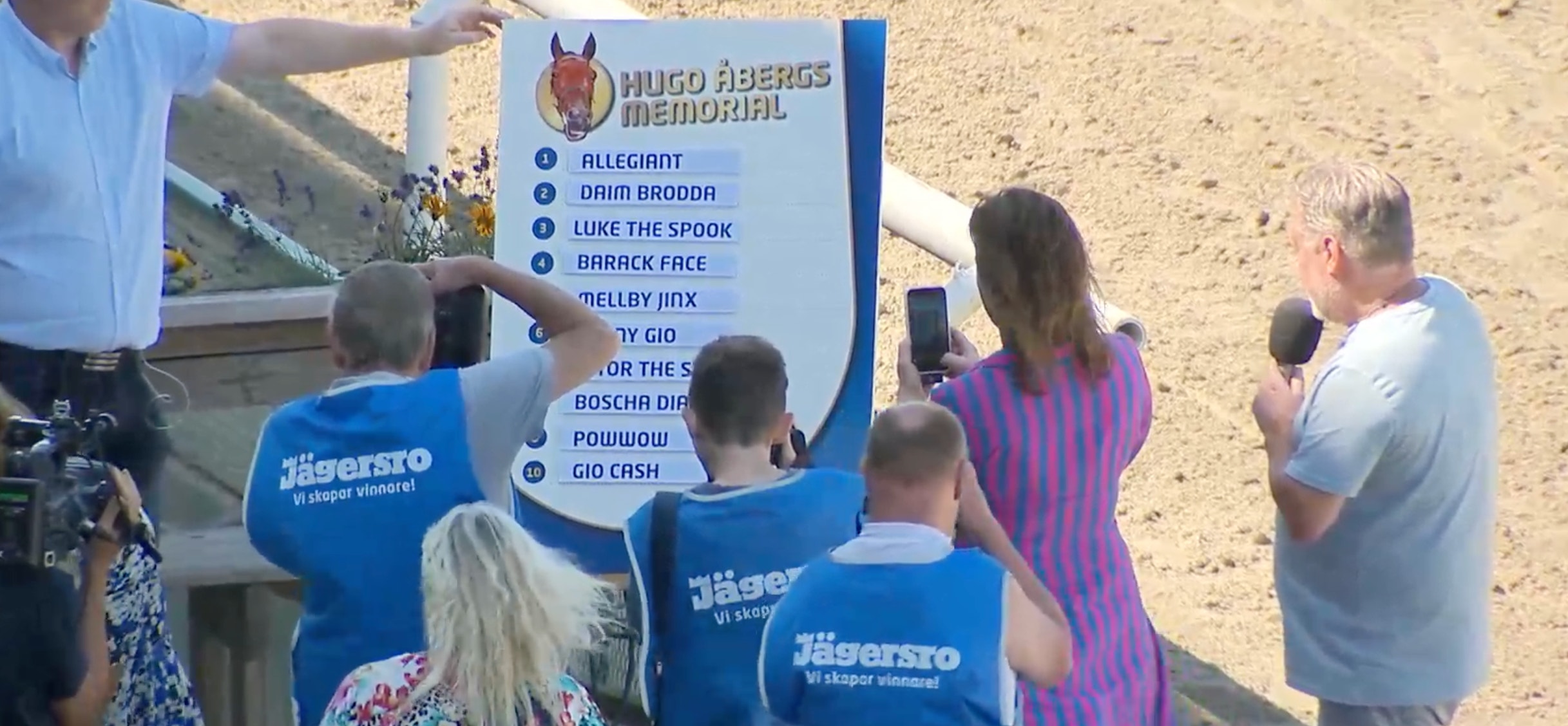 En grupp människor samlade på en sandstrand omkring en blå minnestavla med text. Flera personer bär blå t-shirts med tryck och fotograferar eller läser på skylten.