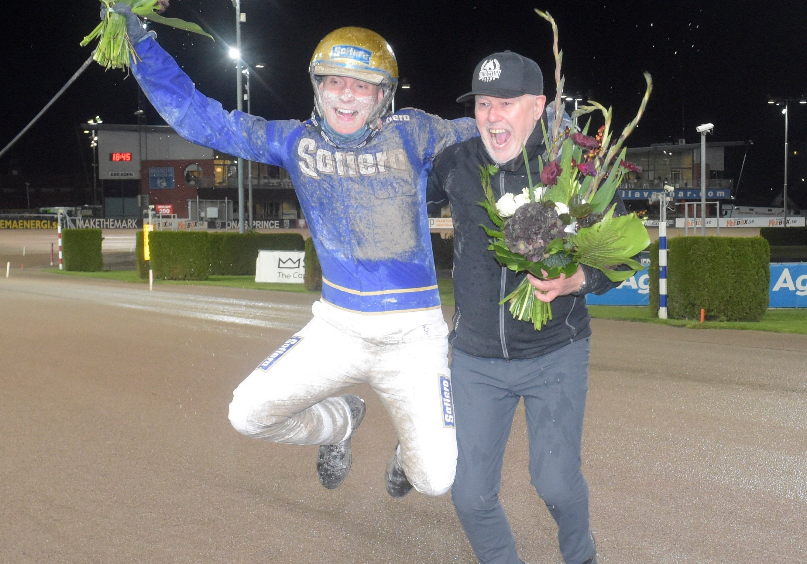 En jockey i blå och vit racingdräkt hoppar av glädje tillsammans med en man som håller en blomsterbukett på en travbana.