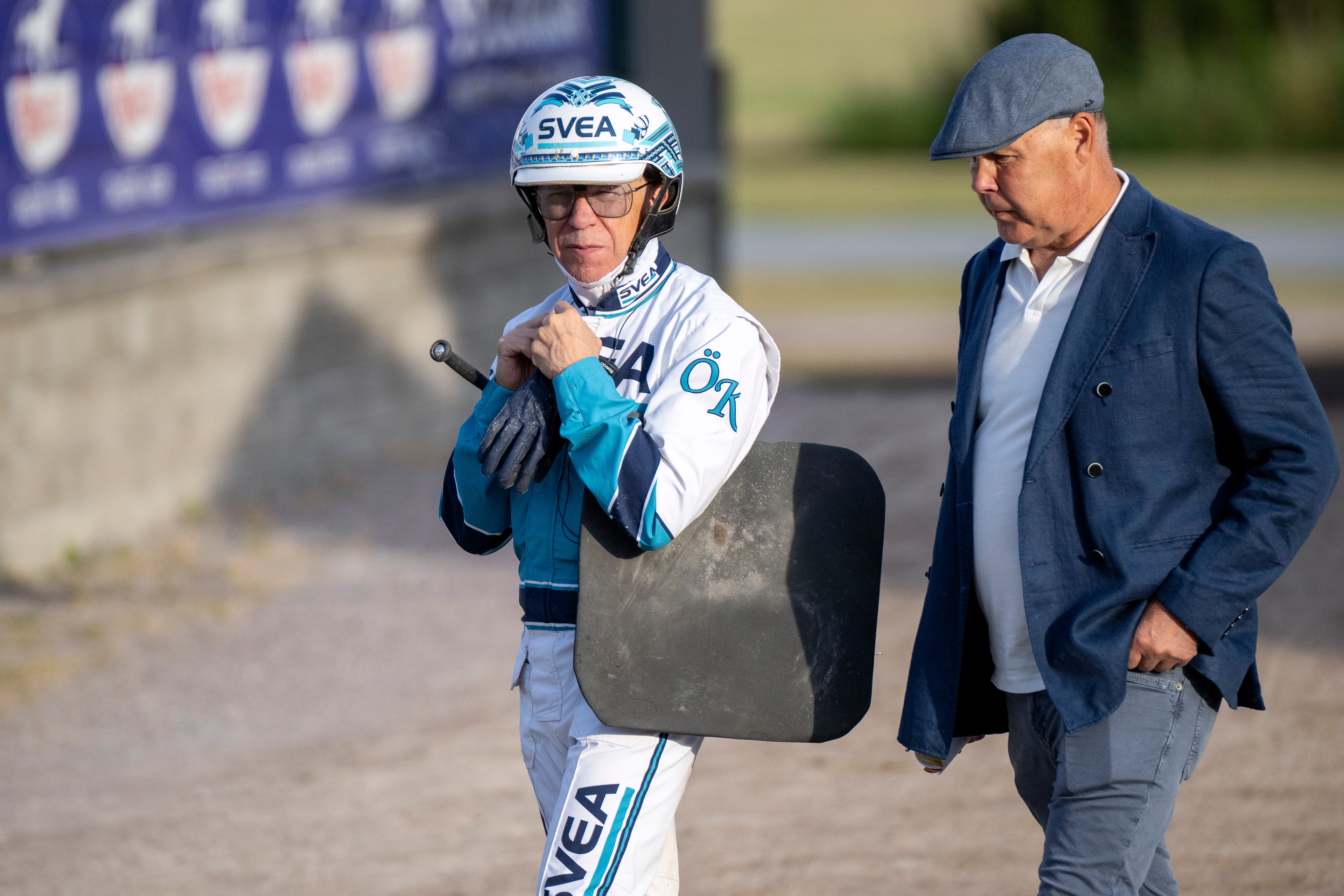 En jockey i vit och blå ridkläder med hjälm och en man i mörk kostym står tillsammans på en travbana.
