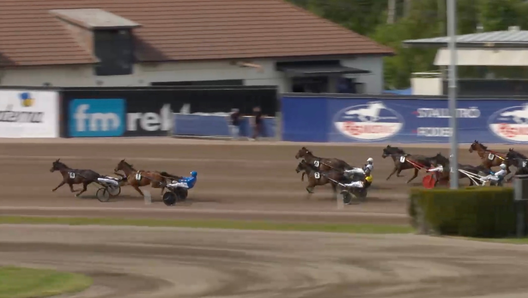 Travlopp med flera hästar som drar vagnar med kuskar på en travbana. I bakgrunden syns tribuntorn och sponsorskyltar.