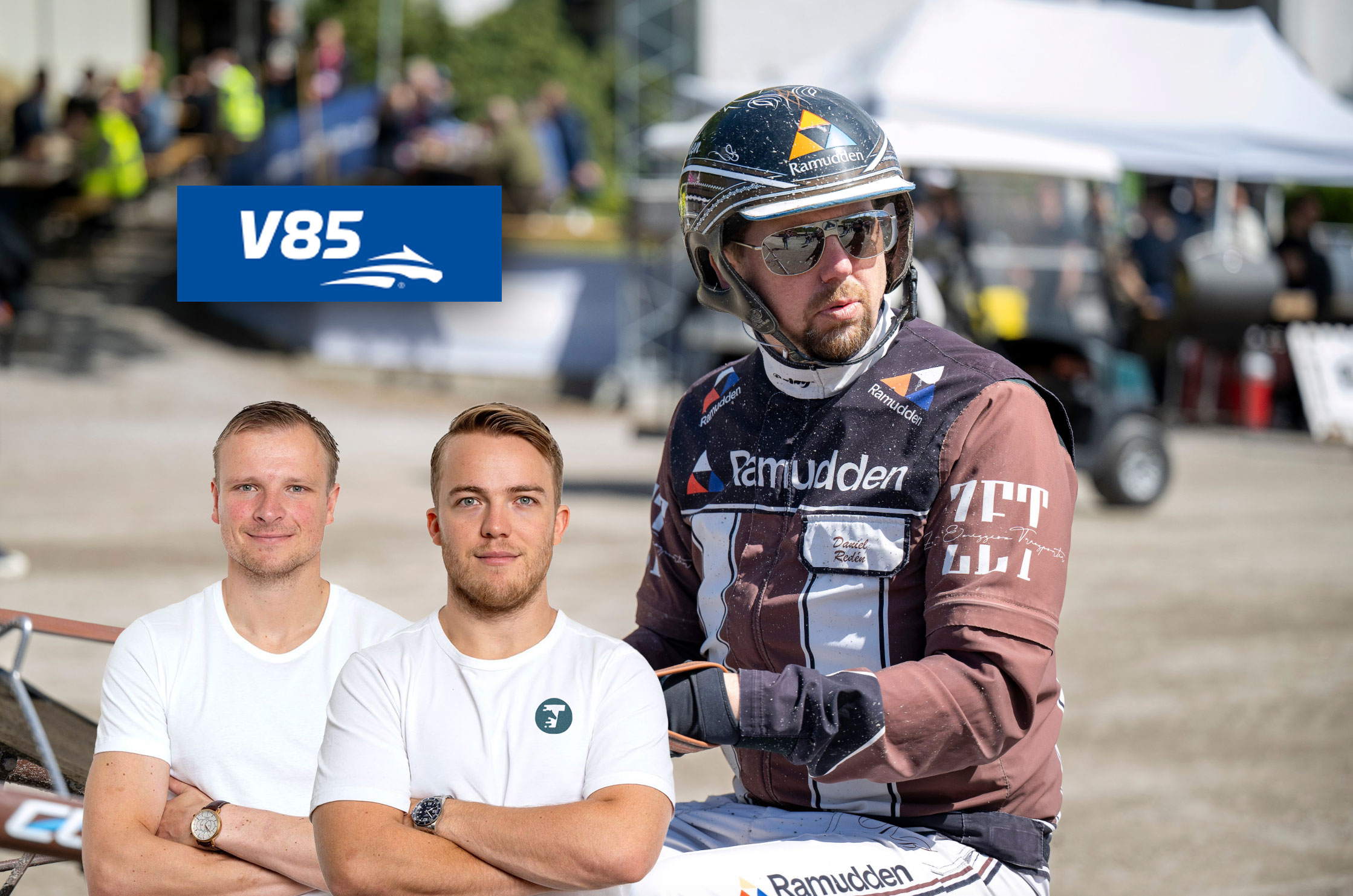 Tre män poserar tillsammans vid ett event. Två män i vita t-shirts står i förgrunden, och en cyklist i cykelkläder och hjälm står bredvid dem. I bakgrunden syns publik och ett tält.
