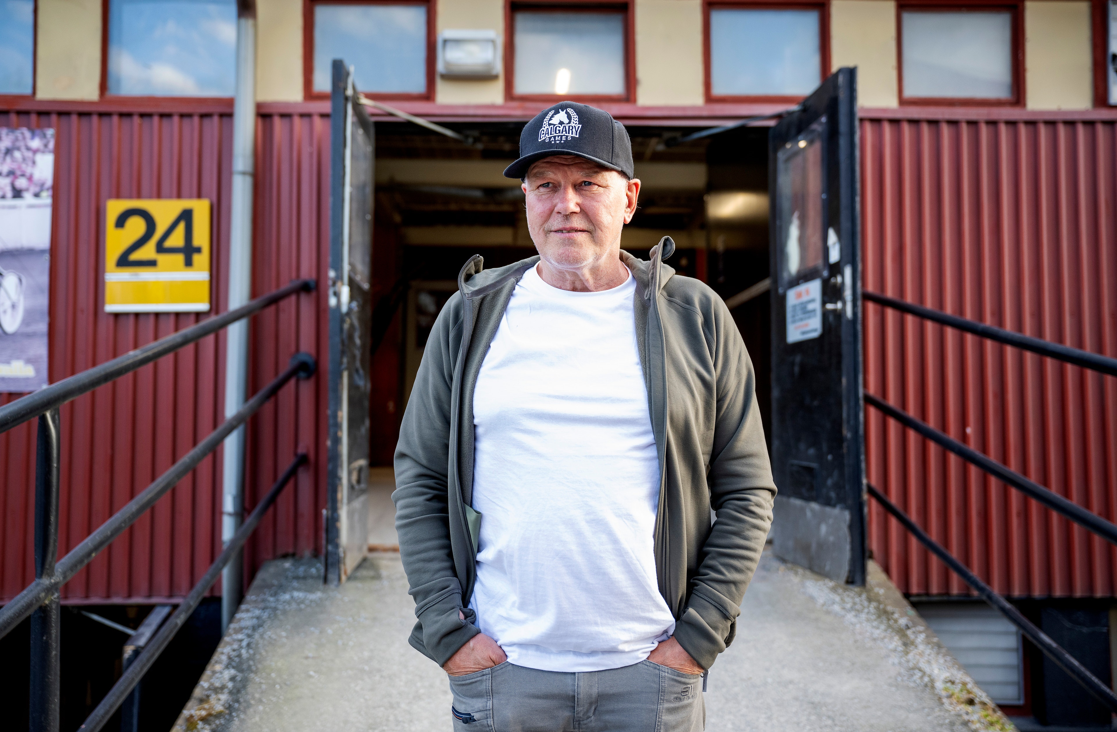 En man står framför en röd metallbyggnad med nummer 24. Han bär basebollkeps, grå hoodie och vit t-shirt.