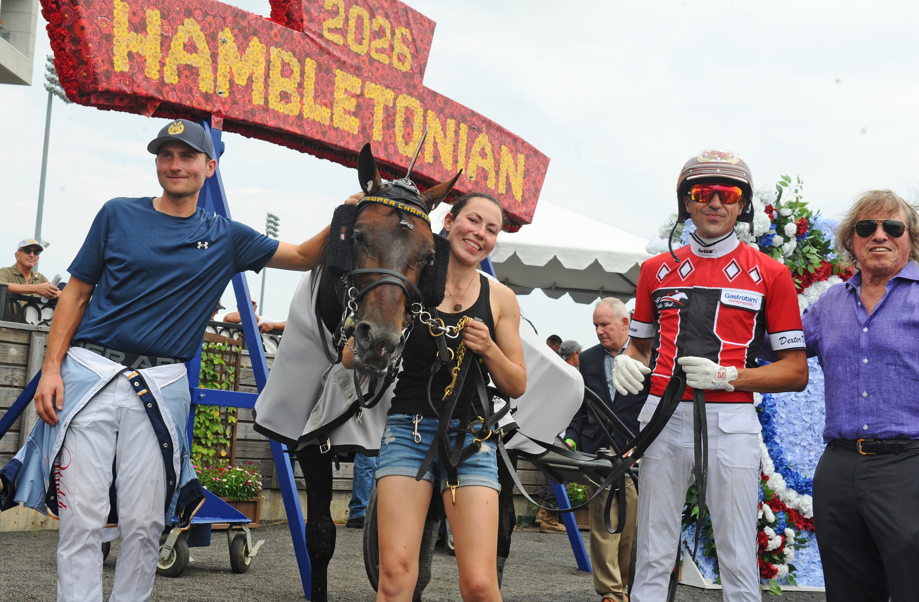 Vinnare och häst presenteras vid Hambletonian-tävlingen. En grupp människor står tillsammans med en häst framför en röd banderoll med texten Hambletonian.