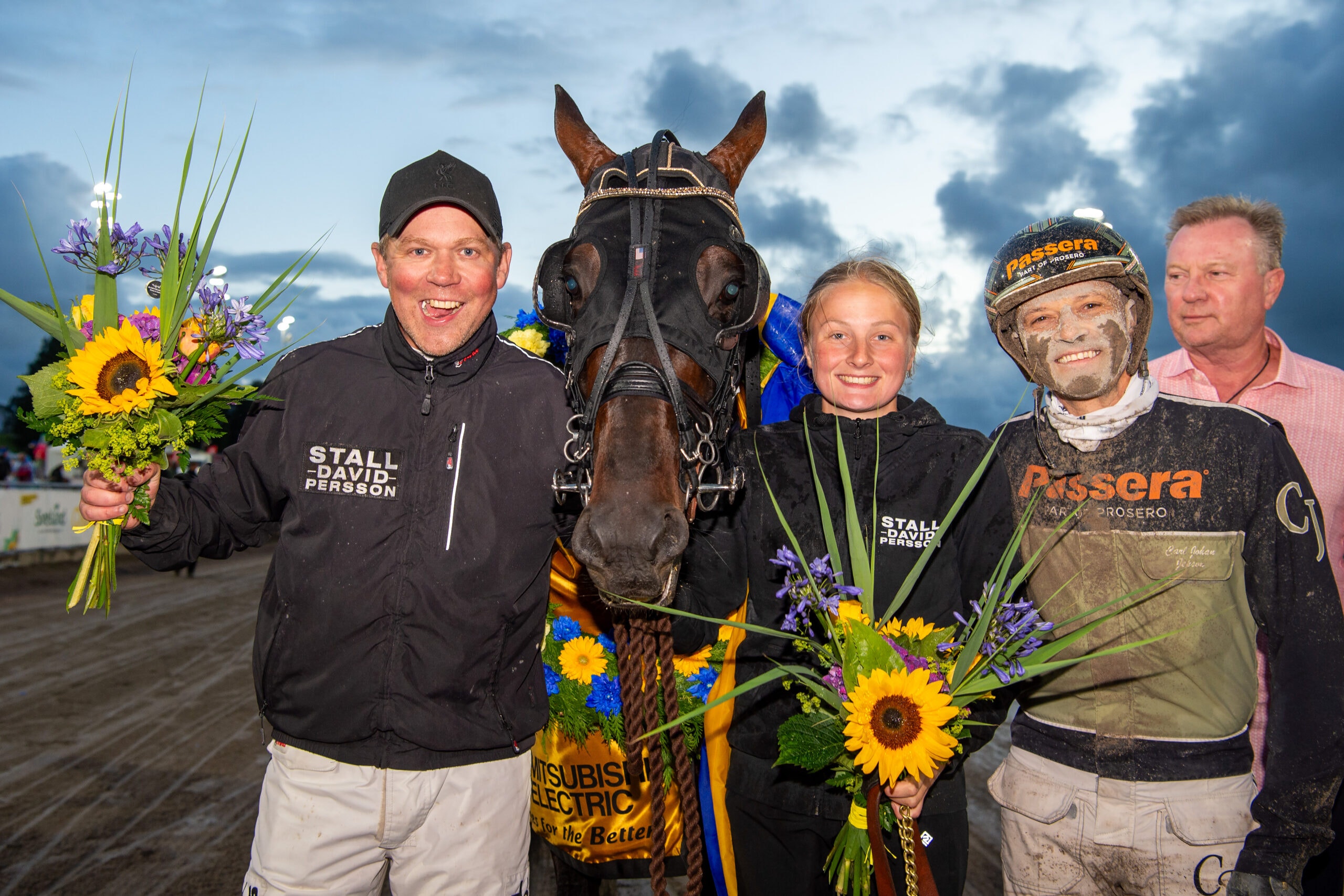 Fyra personer tillsammans med en häst vid en prisutdelning. De håller blommor och ler mot kameran under en molnig himmel.