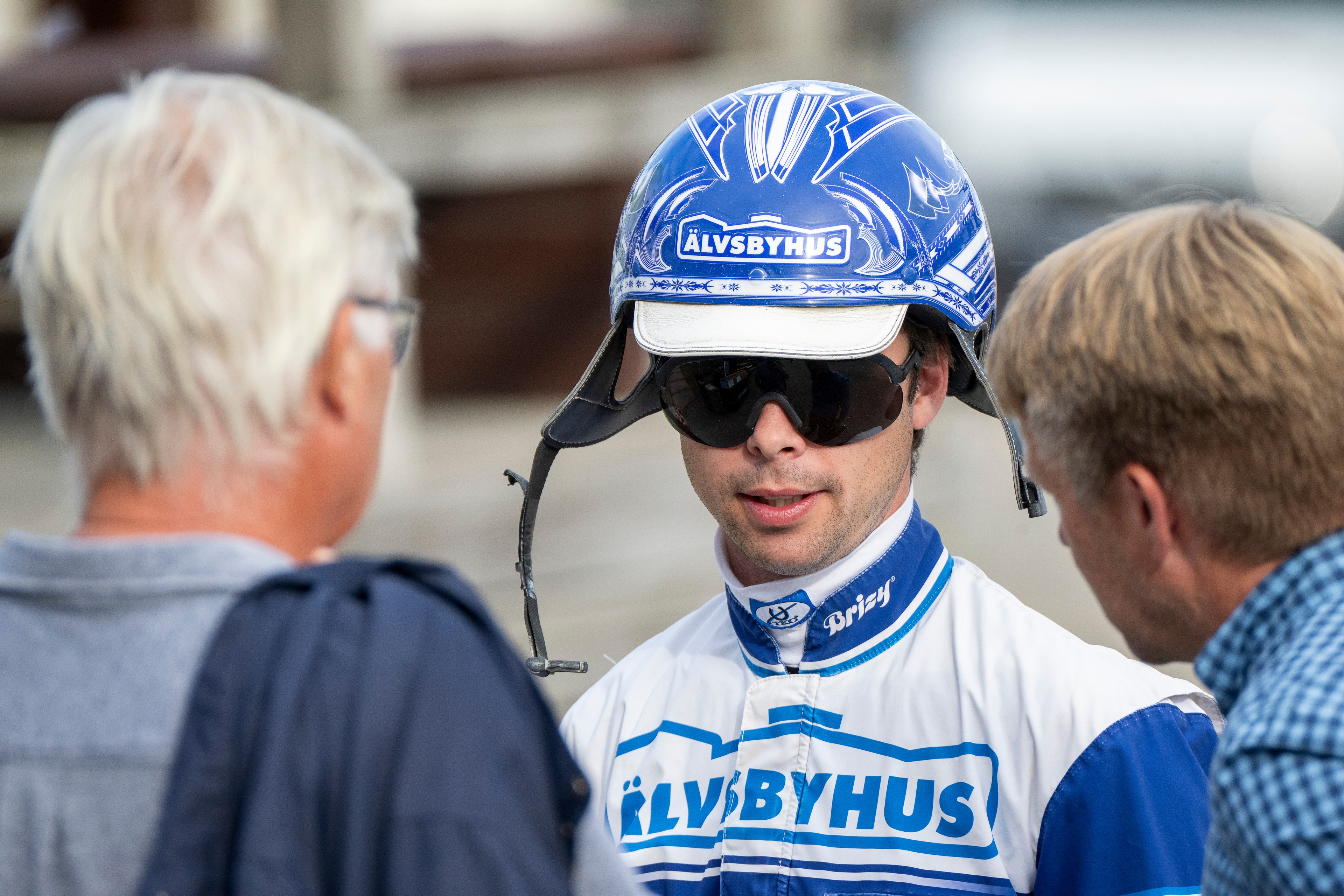 Cyklist i blå och vit träningsdräkt med texten "ELVBYHUS" pratar med två äldre män. Cyklisten bär cykelhjälm och solglasögon.