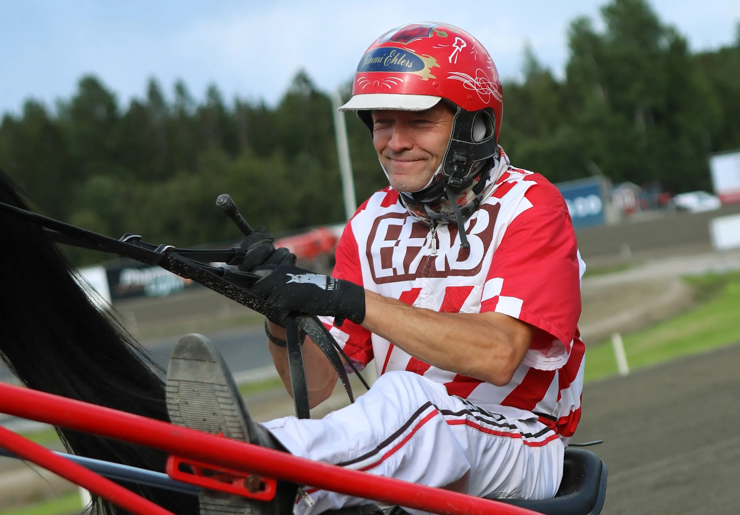 Jimmy Ehlers sitter i en röd sulky på en travbana. Han är klädd i röd och vit sportutrustning med hjälm och handske, och håller i sulkyns tyglar.
