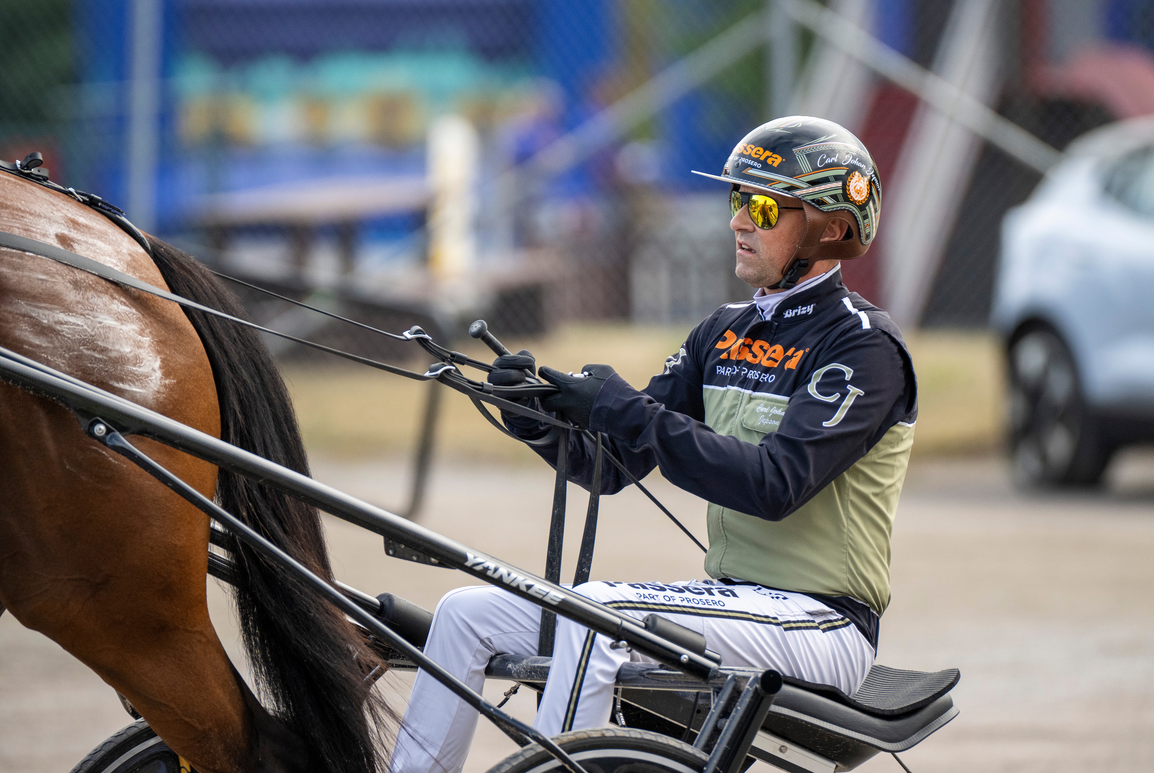 En kuskvagnskusks sittande i en sulky bakom en brun häst. Kusken bär hjälm, gula skyddsglasögon och sportkläder med orange och mörkblå färger. Kusken håller i tyglar till hästen. I bakgrunden syns en parkering och byggnader.