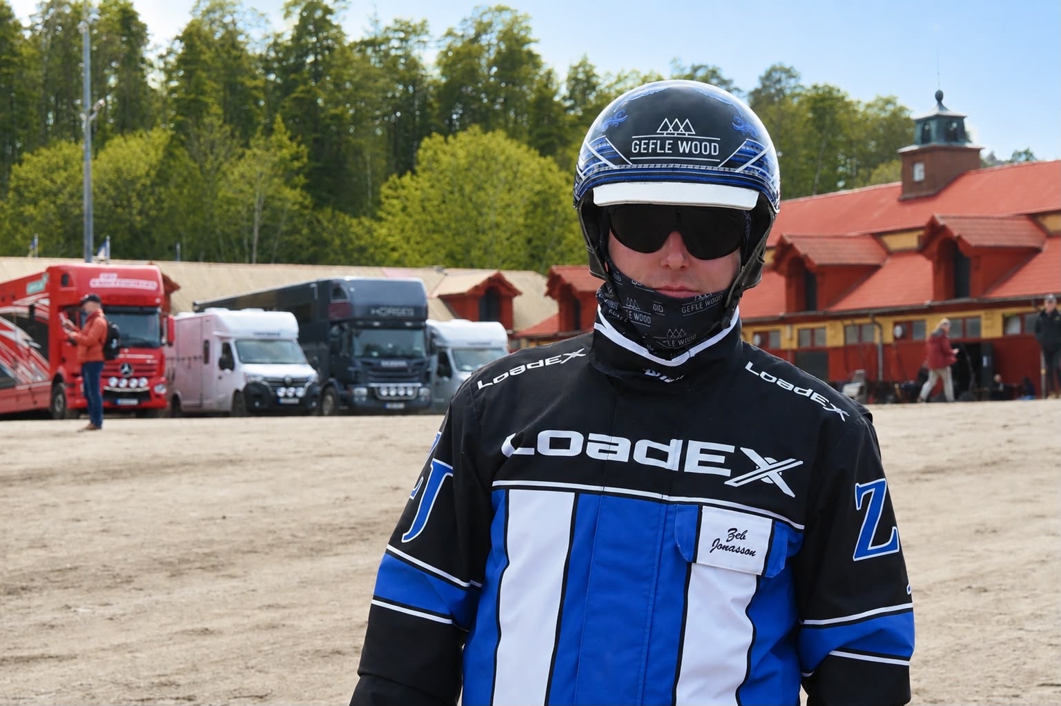 Porträttbild av en person i blå och svart motorsportdräkt med vit hjälm och solglasögon. I bakgrunden syns fordon och en röd byggnad.