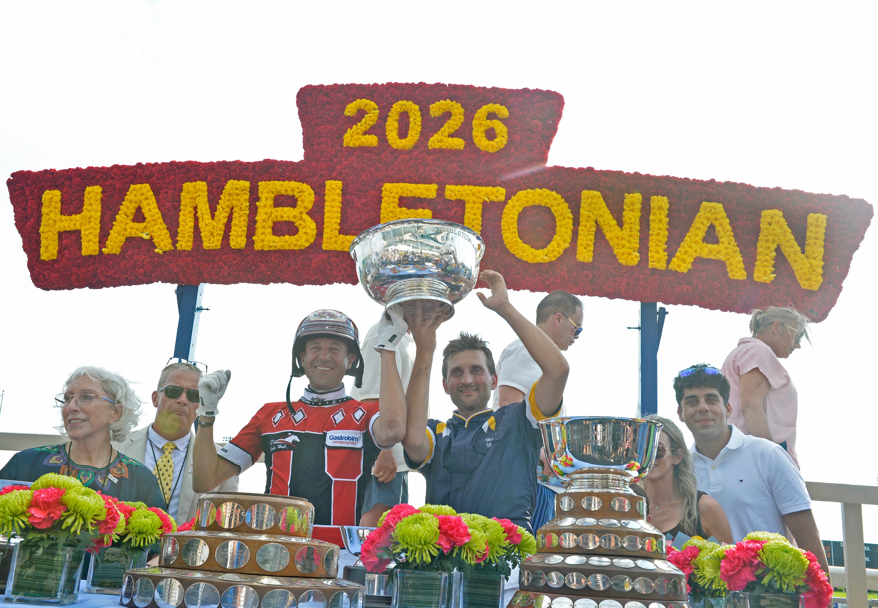 Prisutdelning vid Hambletonian 2026. Vinnare och officiella personer står med pokaler och blommor under en brun skylt med gul text.