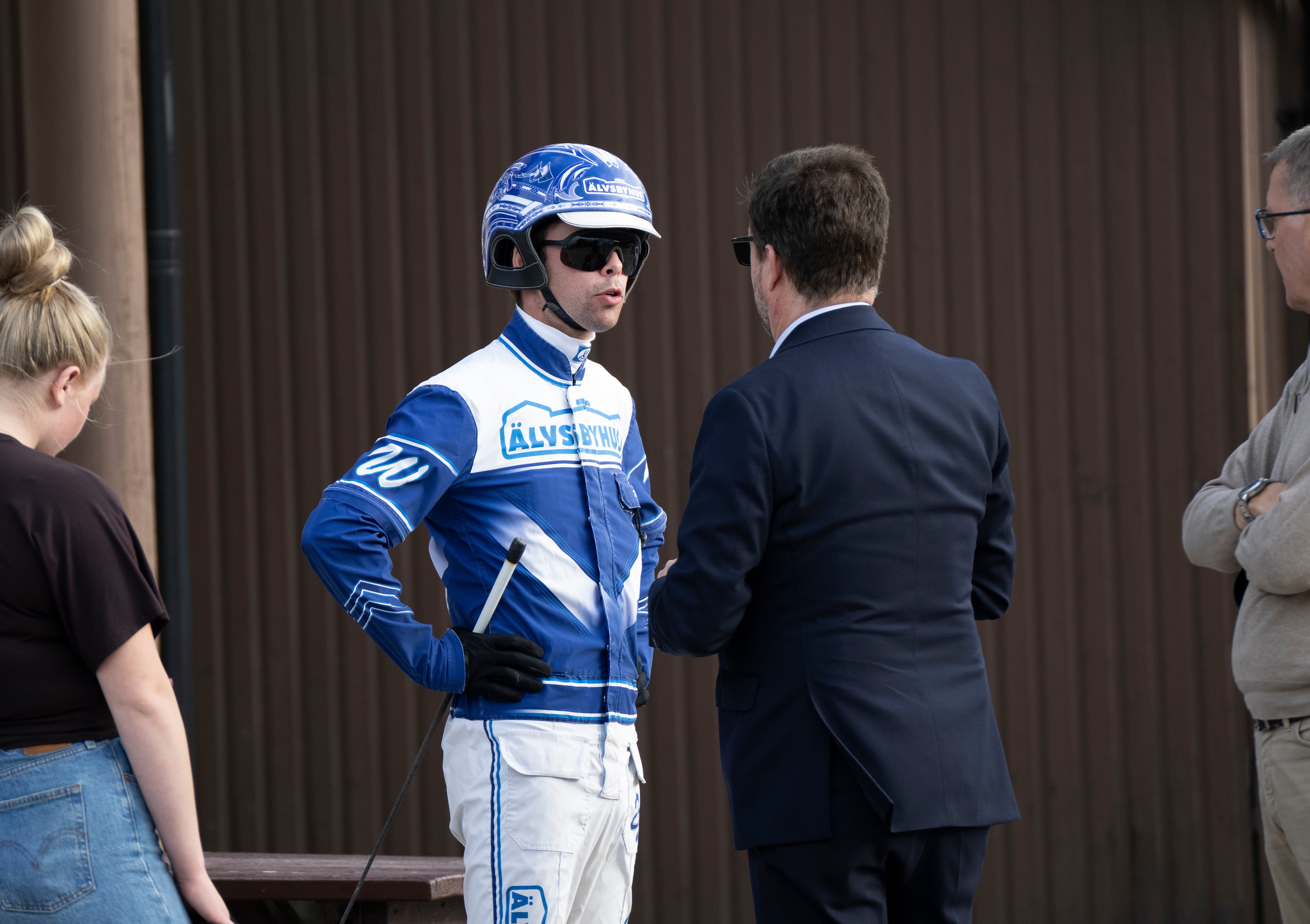 En jockey i blå och vit racing-dräkt med hjälm och solglasögon står och samtalar med personer vid sidan av en bana. Daniel Wäjersten