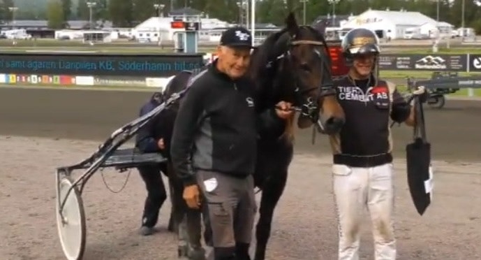 Två personer leder en mörkbrun häst med sulky på en travbana.