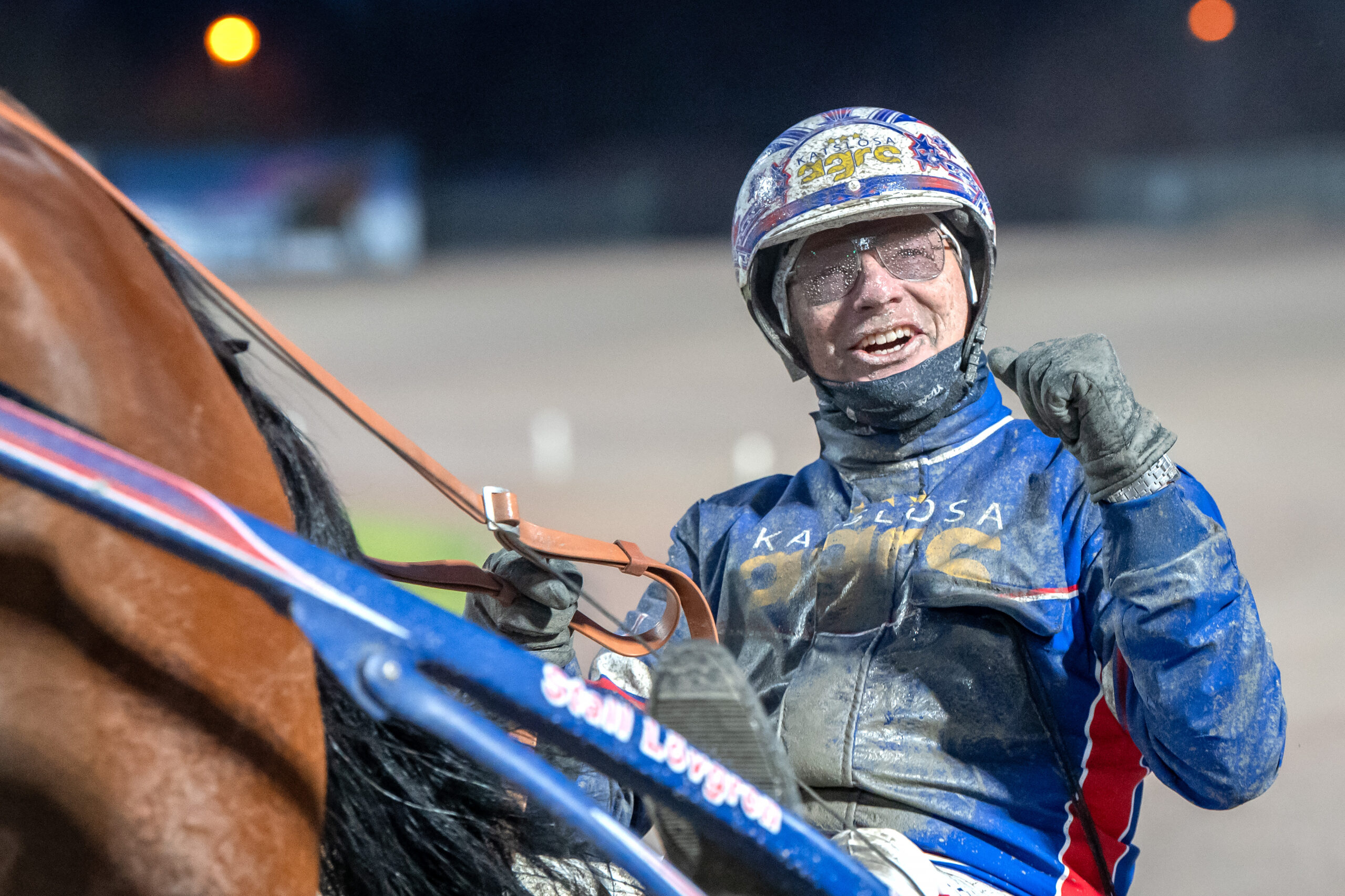 Porträtt av en jockey i blå racing-outfit och hjälm som håller i tygeln på en häst under kvällen.