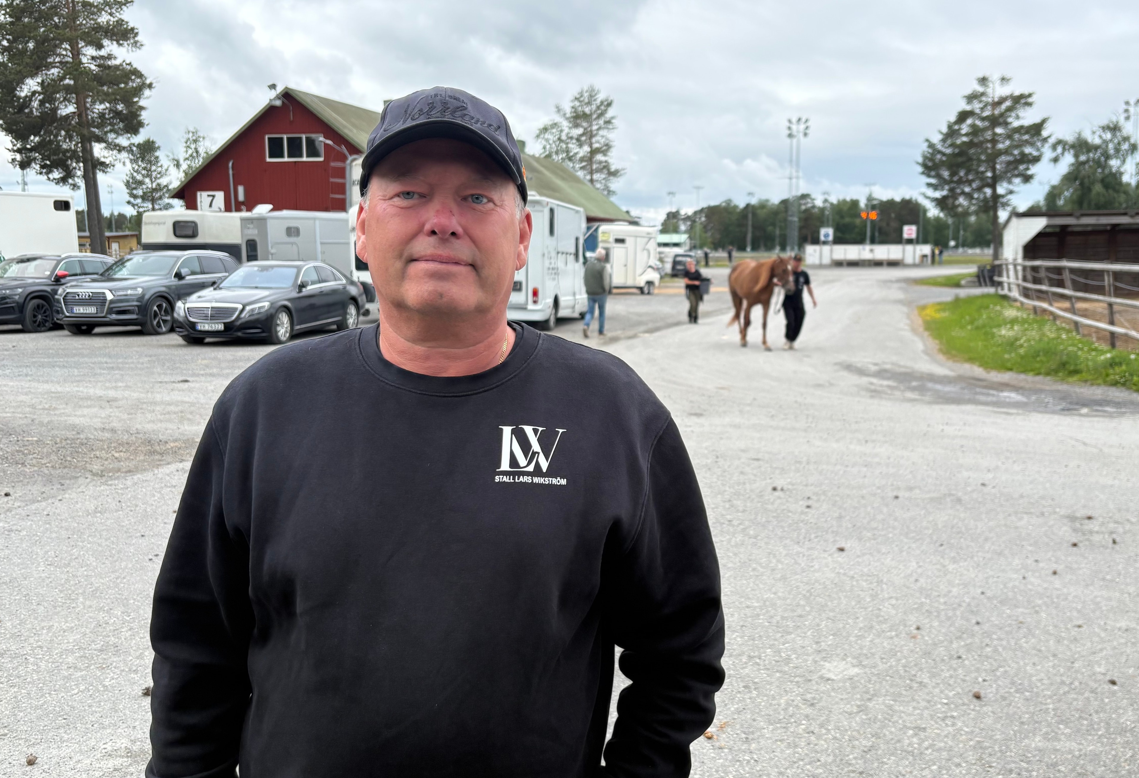 Porträtt av en man i svart tröja och mörkblå keps som står utomhus på en lantgård. I bakgrunden syns röda och vita byggnader, parkerade bilar och hästar.