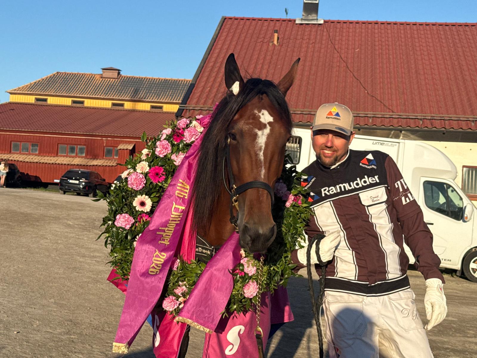 En man i racing-outfit står bredvid en brun häst som är utsmyckad med en rosa täcke och en blomsterkrans. I bakgrunden syns röda stall och byggnader.