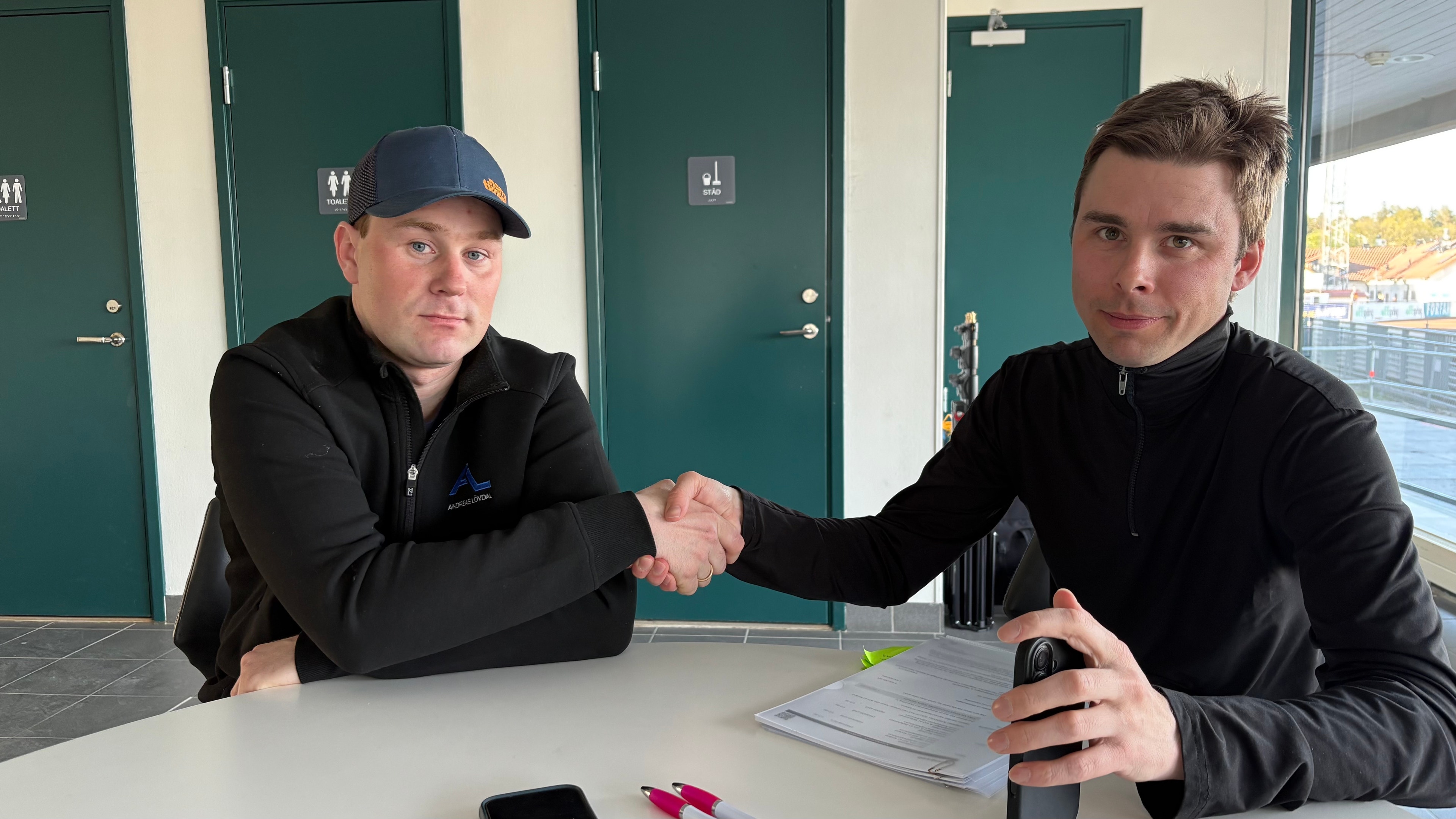 Två män skakar hand vid ett bord. En man i svart jacka och keps på vänster sida, en man i svart jacka på höger sida. I bakgrunden gröna dörrar och ett fönster.
Foto: Patrick Sjöö