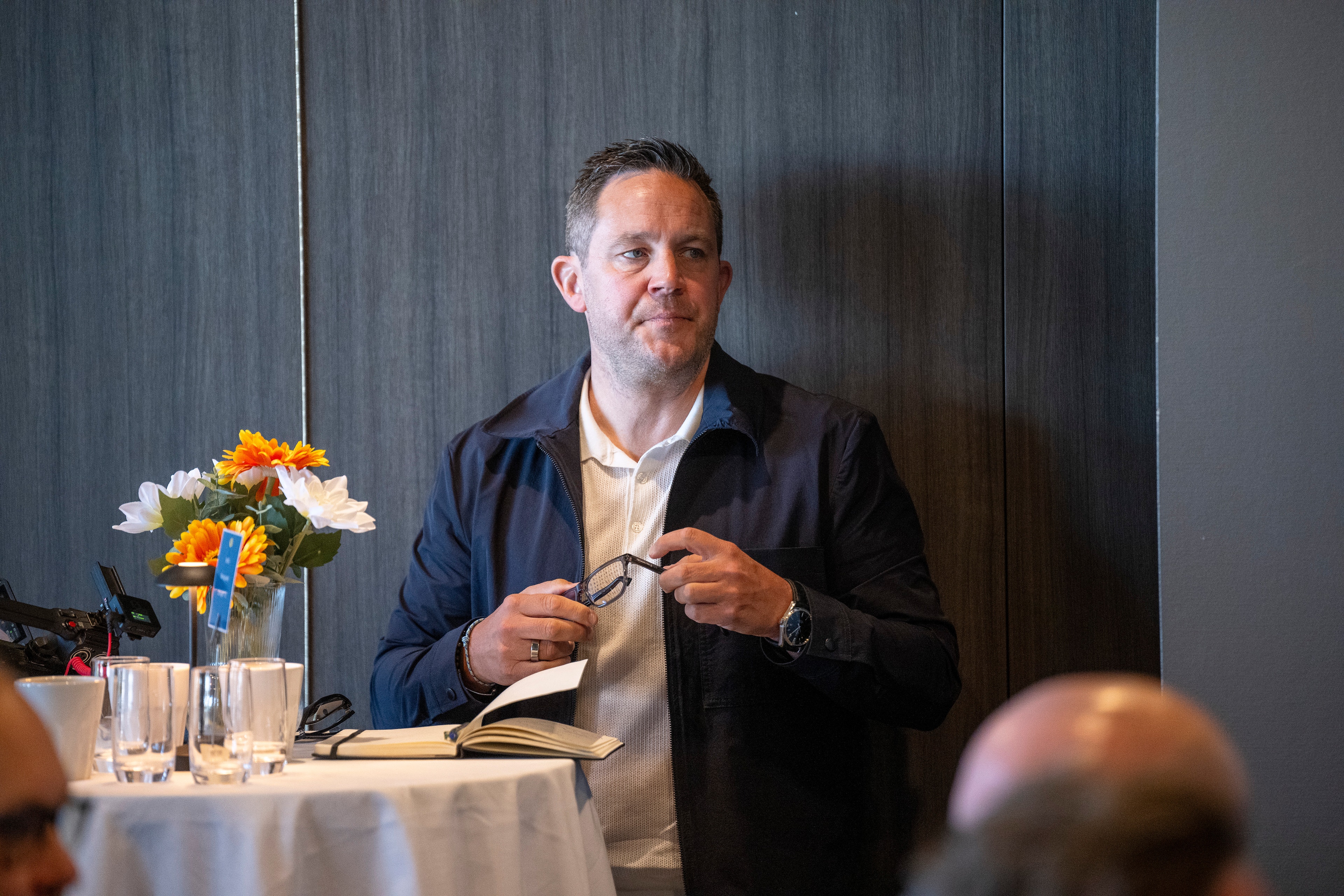 En man i mörkblå kavaj står vid ett bord under en presentation. Han håller i en anteckningsbok och framför honom står en vas med gula och vita blommor.