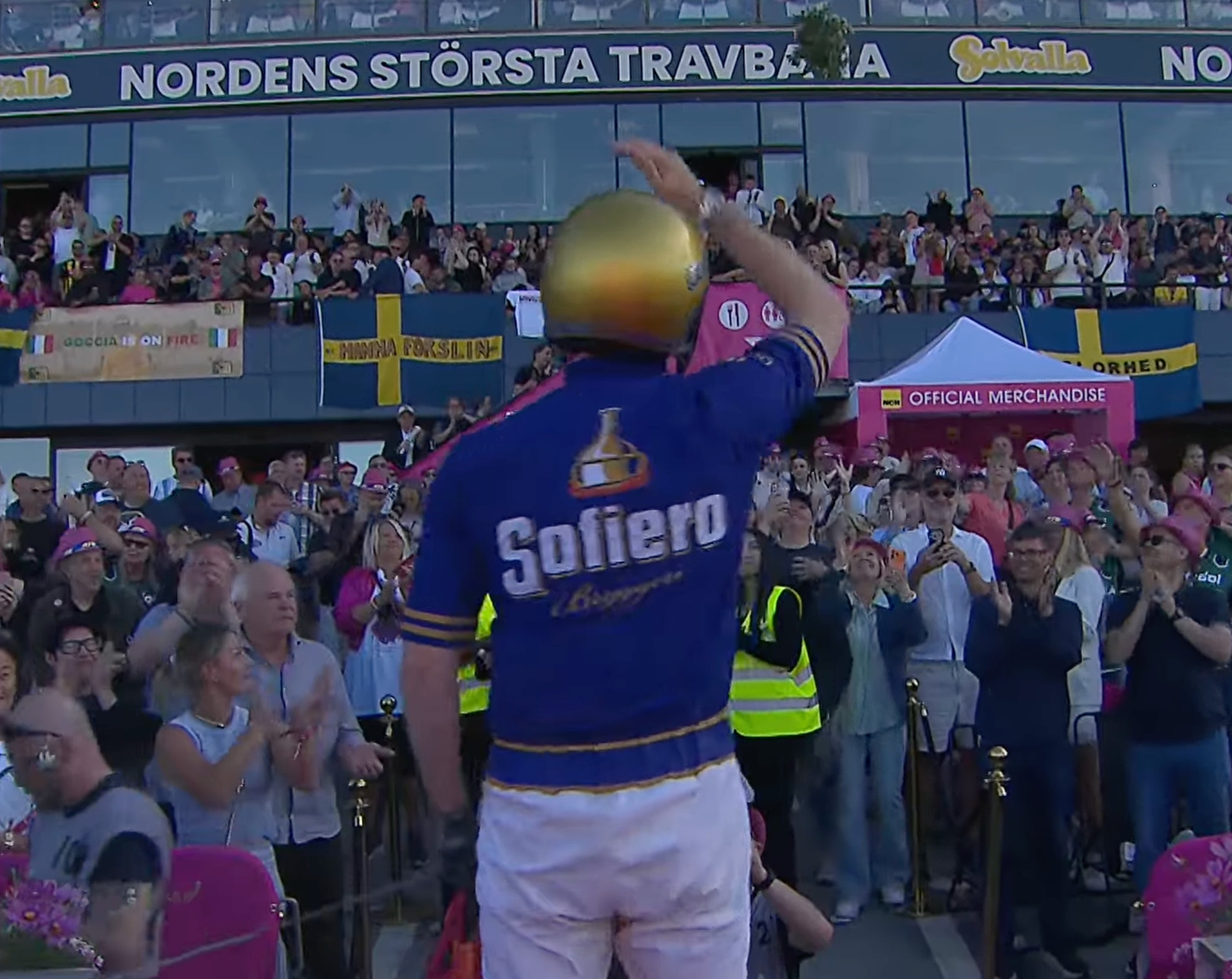 Fotbollsspelare i blå och rosa tröja håller upp en gul boll framför en fullsatt fotbollsstadion med publik.