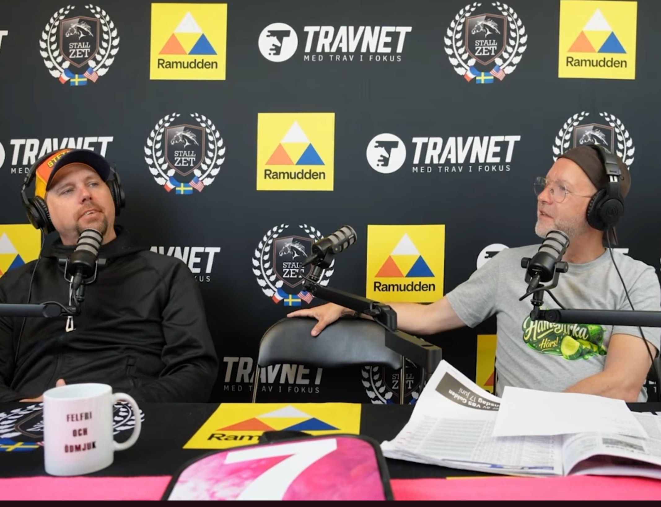 Två män sitter vid ett podcastbord med mikrofoner och headset framför en sponsorvägg med logotyper för Travnet och Ramudden.