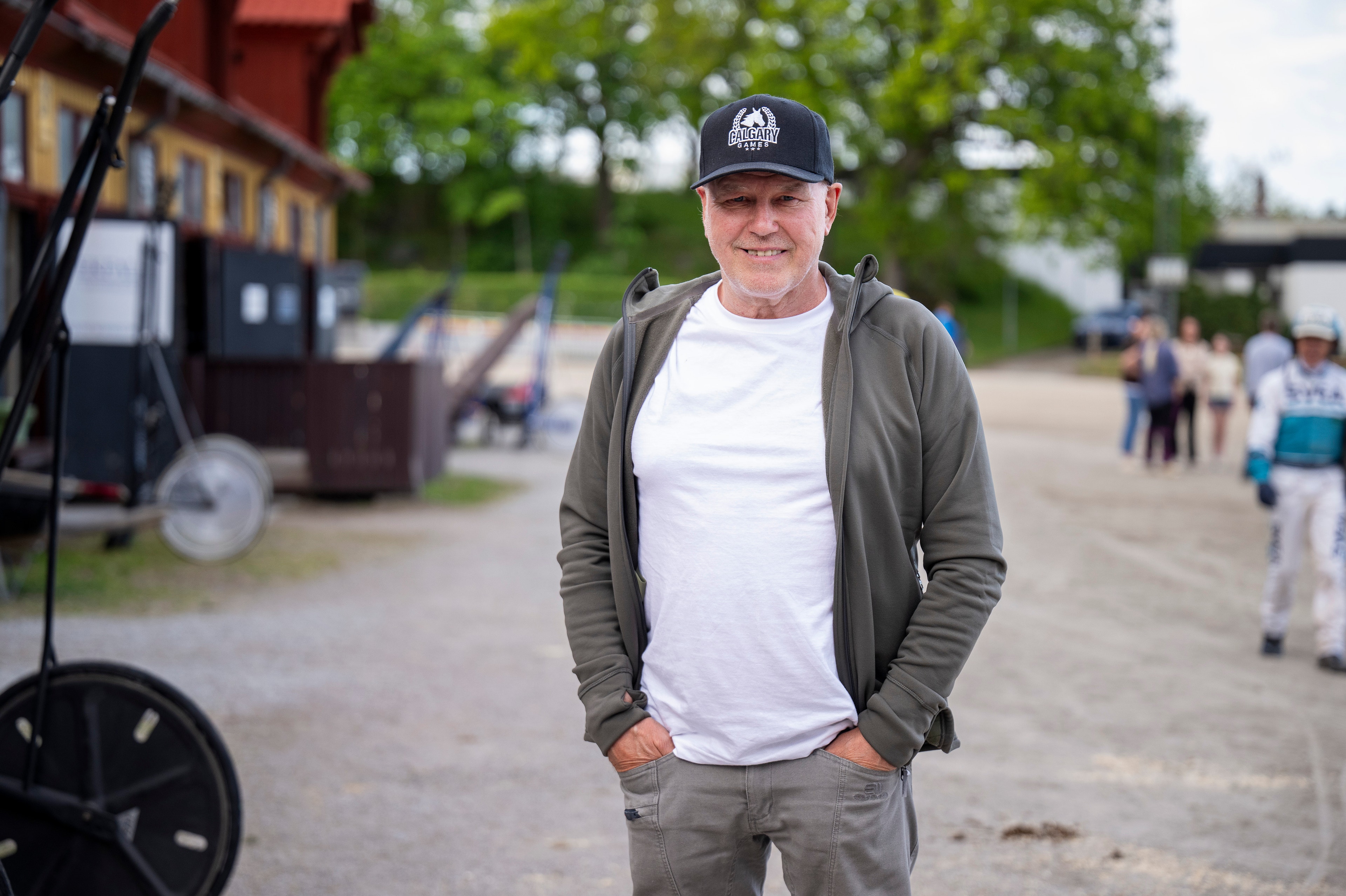 Porträtt av en man som står på en öppen plats. Han bär en vit t-shirt under en grå hoodie och en svart keps.