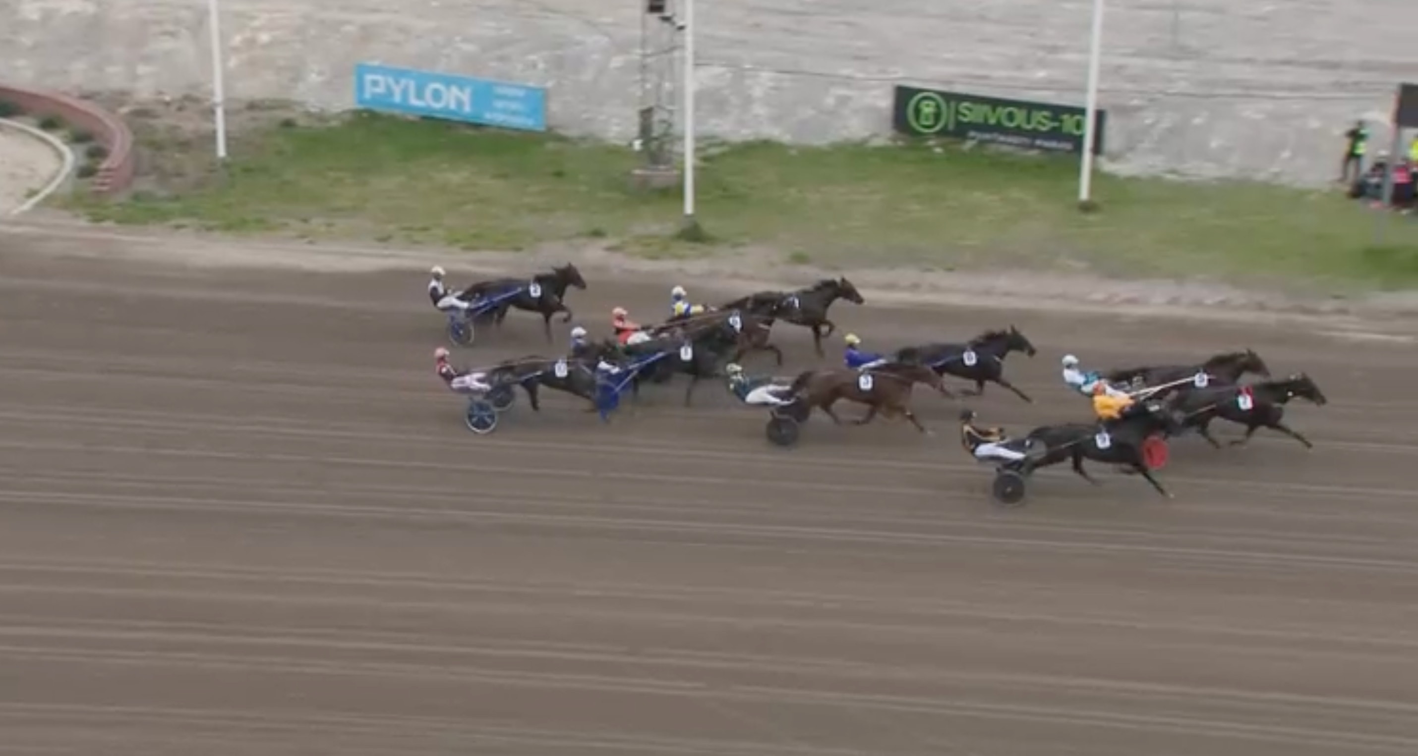 Luftfoto av ett travlopp med flera hästar och kuskar som springer på en sandtrack under tävling.