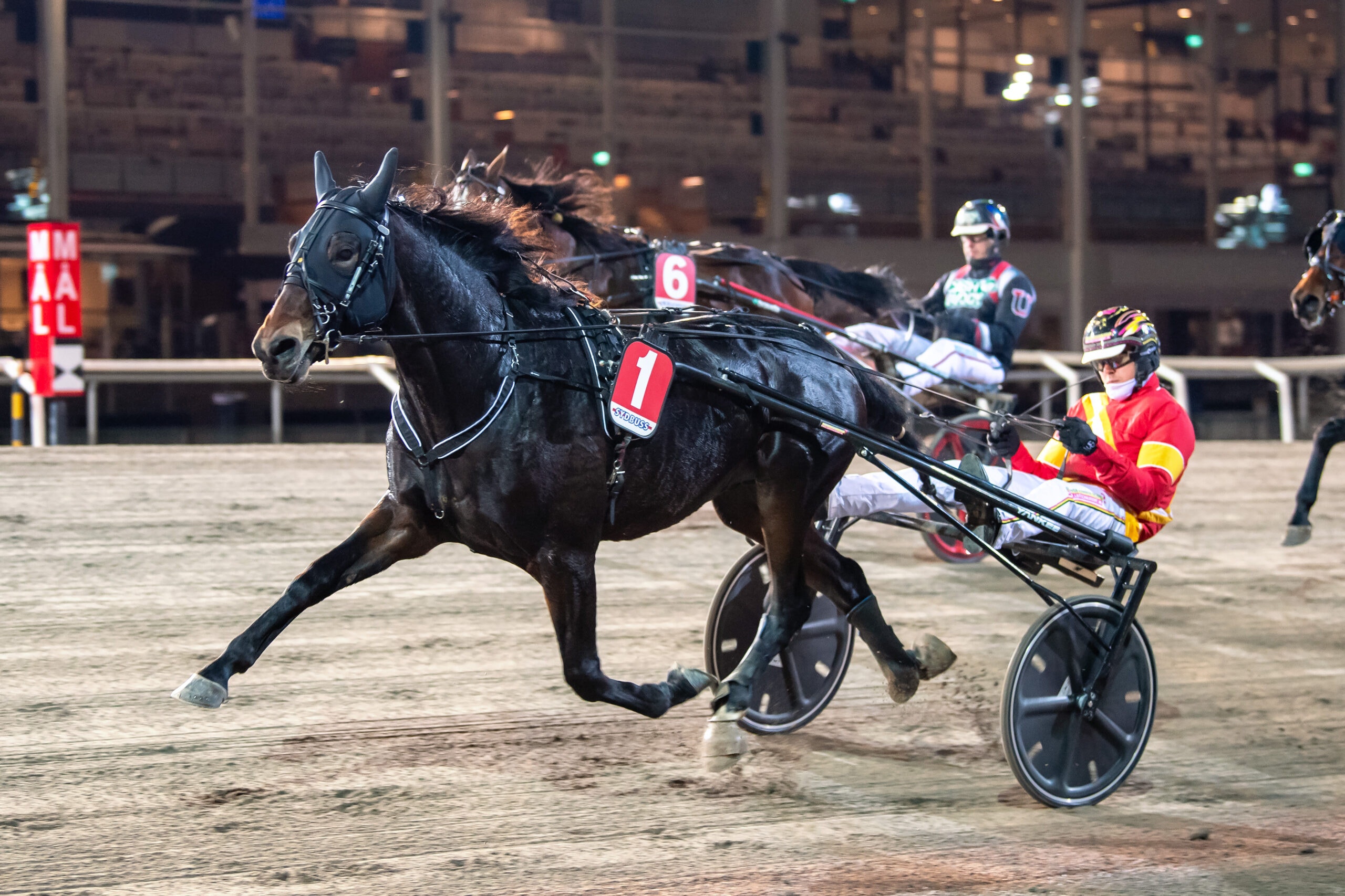 En svartvit häst i full galopp drar en lätt sulky med en kuskar i gul och röd tävlingsdräkt under ett travlopp på en sandtrack.